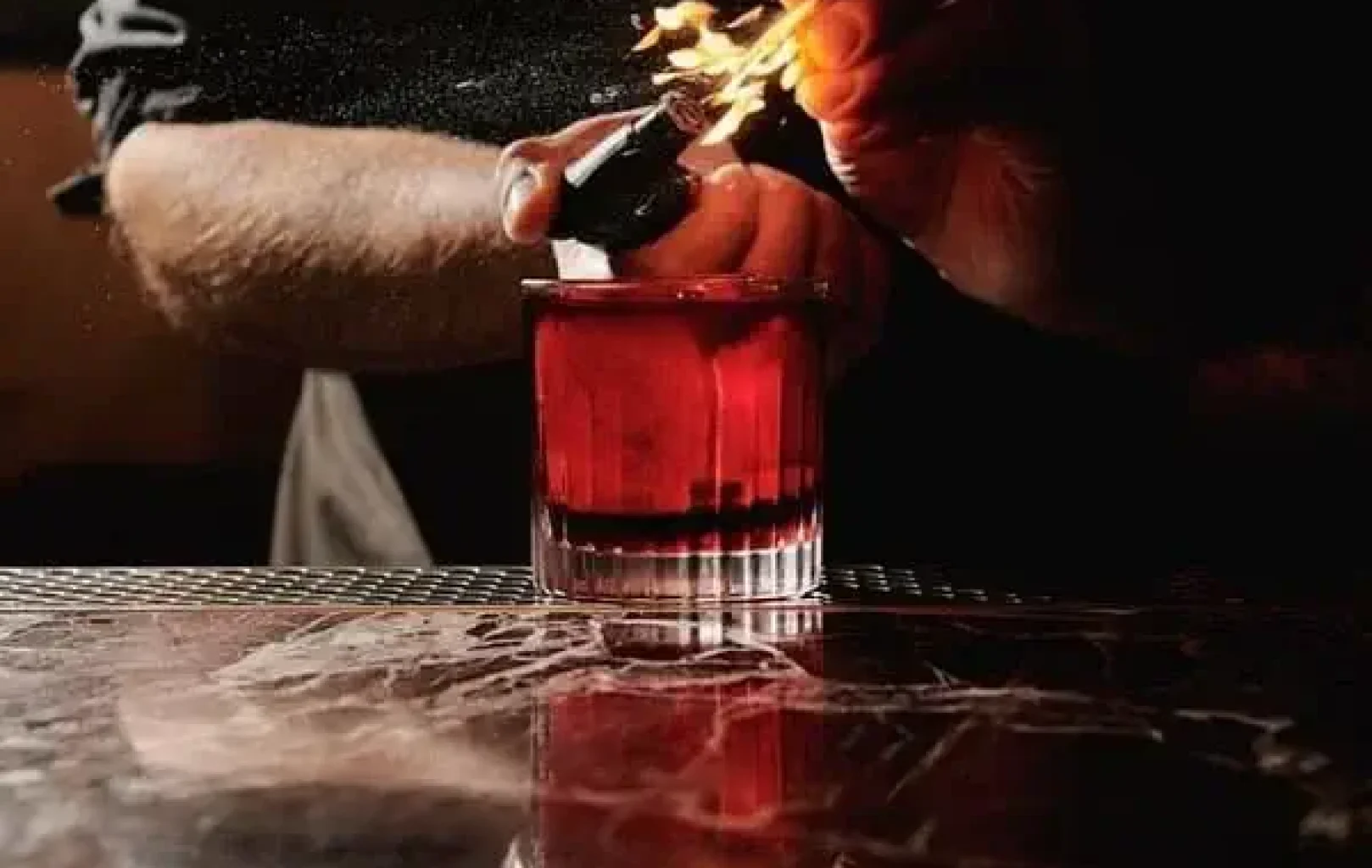 Eine Person hält ein Feuerzeug, das eine Flamme über einem roten Cocktail in einem Glas auf einem Bartresen entzündet.