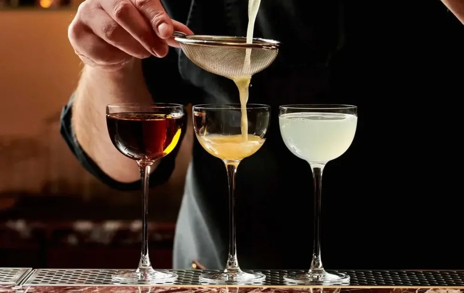 A person strains a yellow cocktail into a coupe glass, with two other drinks—one dark and one light—already poured and lined up on a bar, ready for Signature Tours to showcase their mixology highlights.