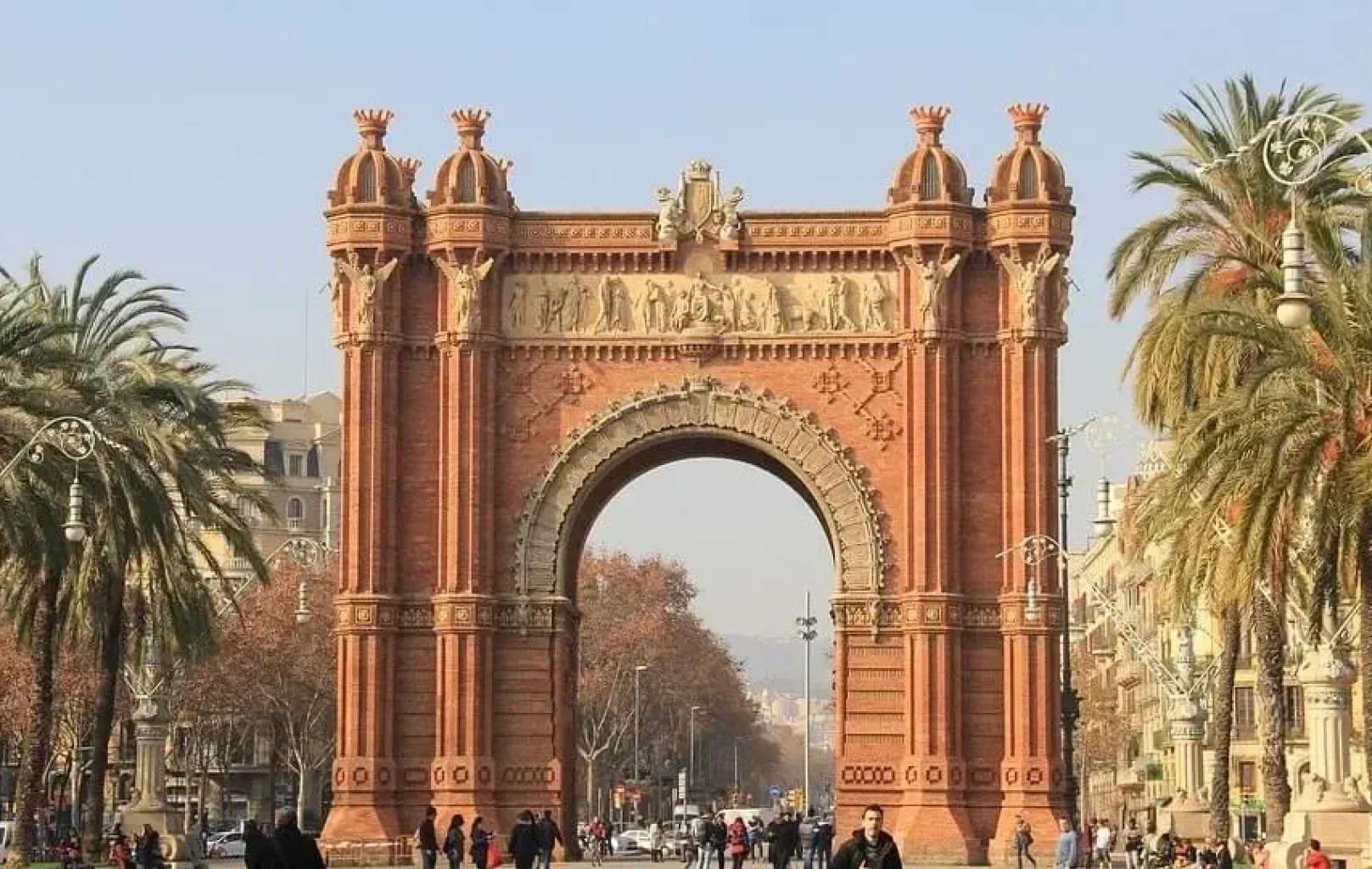 Un gran monument d'arc de maó vermell, l'Arc de Triomf de Barcelona, es troba al final d'un ampli passeig de vianants vorejat de palmeres i gent gaudint dels dies assolellats de març o abril.