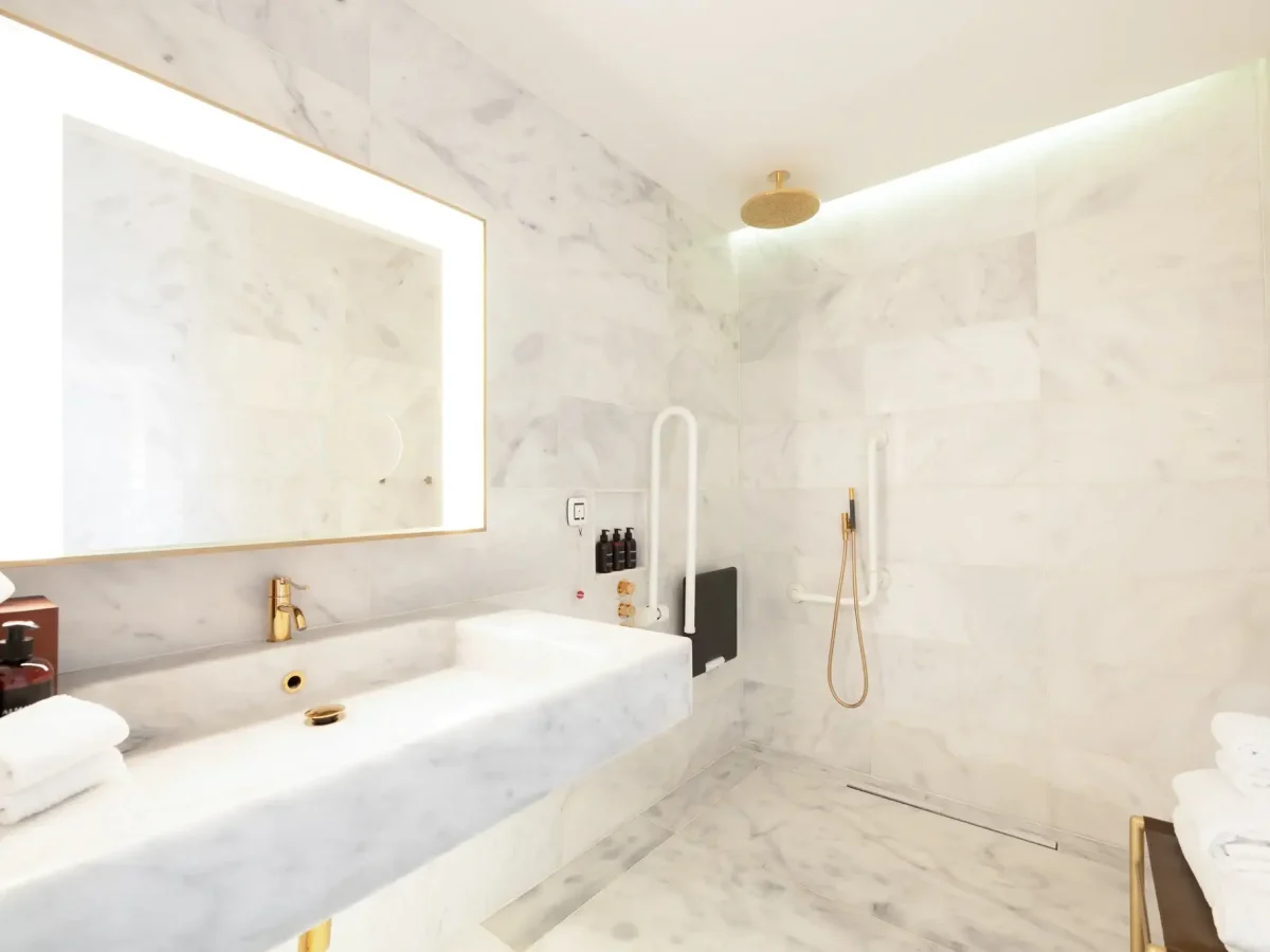 Moderno cuarto de baño accesible con paredes y lavabo de mármol, un gran espejo iluminado, accesorios dorados, ducha a ras de suelo, barras de sujeción y toallas plegadas: perfecto para una habitación adaptada que ofrece estilo y funcionalidad.