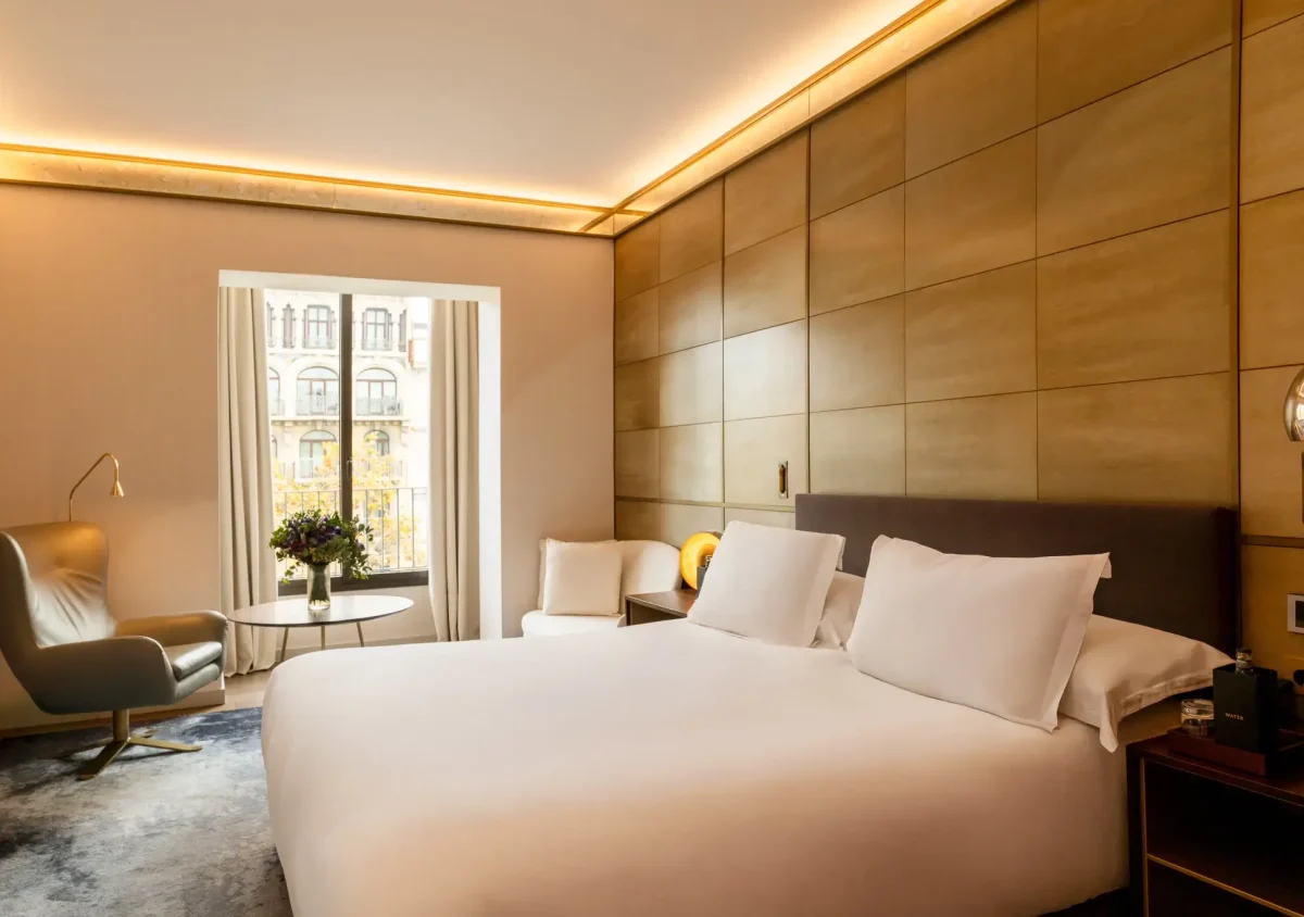 Ein modernes Hotelzimmer von Almanac Zimmer mit einem großen Bett, zwei weißen Stühlen, einem runden Tisch mit Blumen, einem grauen Sessel und einem Fenster mit Blick auf eine belebte Straße in der Stadt.