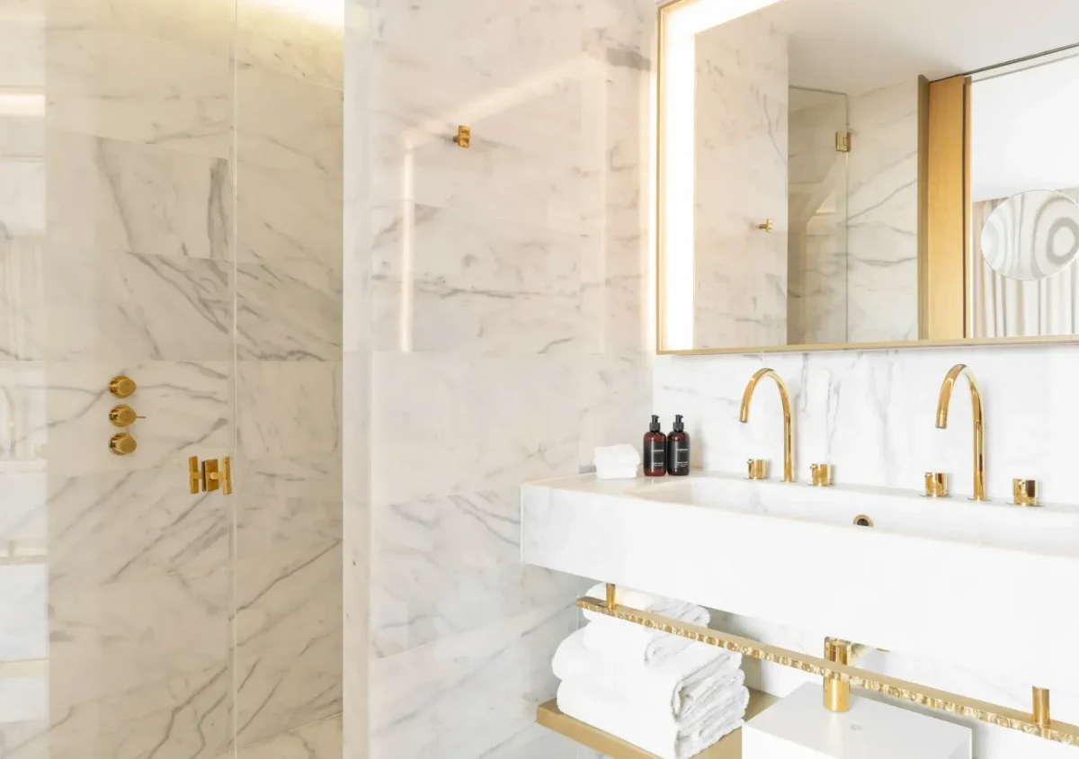 Modernes Badezimmer mit weißen Marmorwänden, goldenen Armaturen, einer Glasdusche, einem Doppelwaschbecken mit goldenen Armaturen und einem großen Spiegel - perfekt für ein Almanaczimmer oder eine Suite mit Balkonzugang. Ordentlich gestapelte weiße Handtücher liegen auf der Ablage.