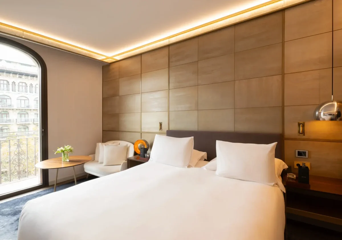 Modernes Almanaczimmer mit einem großen Bett, weißem Bettzeug, zwei Nachttischen, einem kleinen Tisch mit Blumen, einem Sessel und einem großen Bogenfenster mit Blick auf die Stadt und Zugang zu einem privaten Balkon.