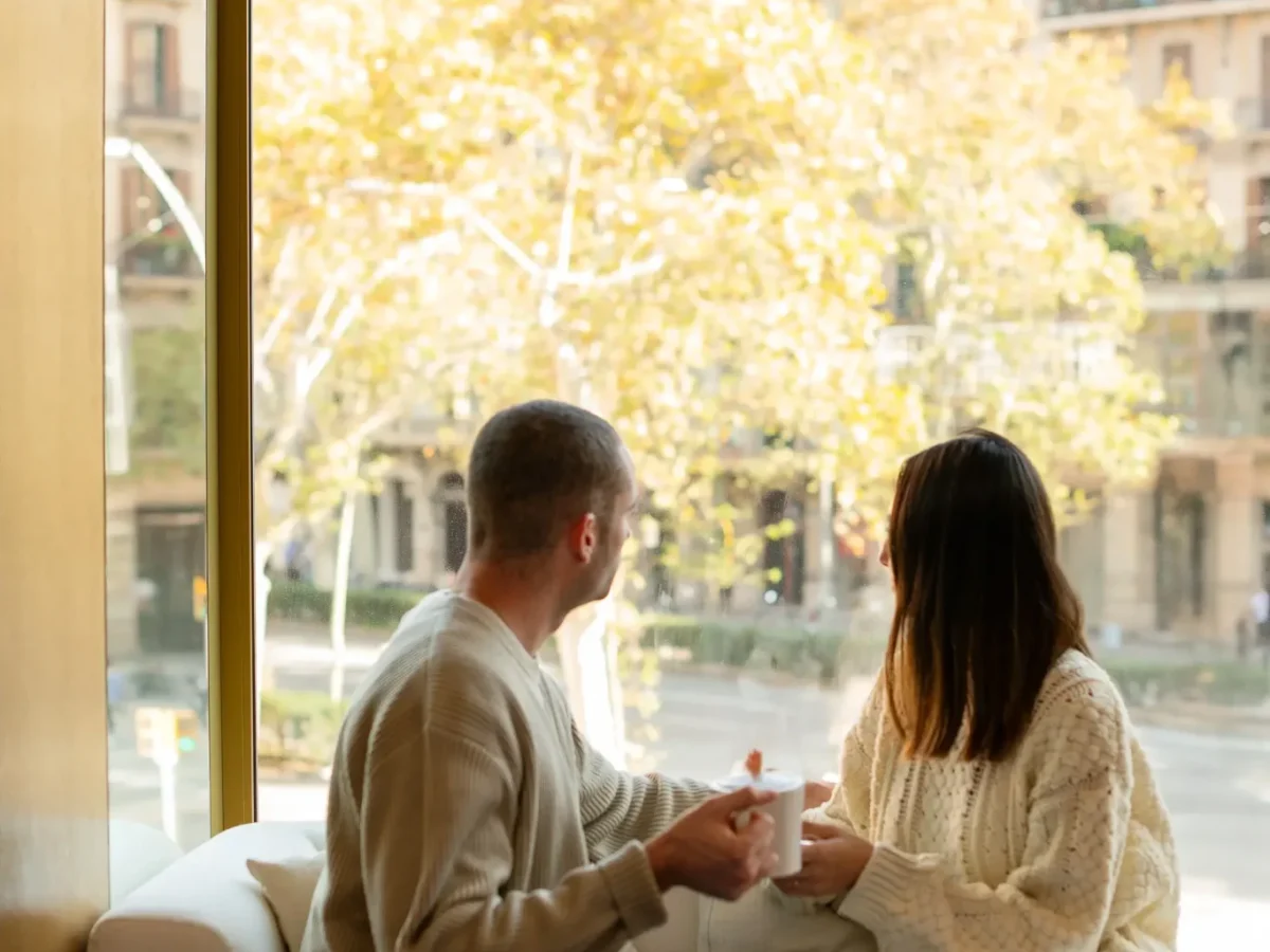 Two people sit on a couch in the Almanac Room by a large window, holding mugs and gazing out at a cityscape of trees and buildings on a sunny day.