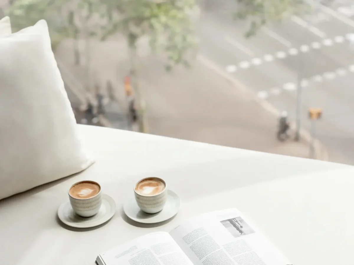 Two cups of coffee and an open book rest on a white surface in the Almanac Room by a large window, overlooking a street intersection with blurred trees and traffic outside.