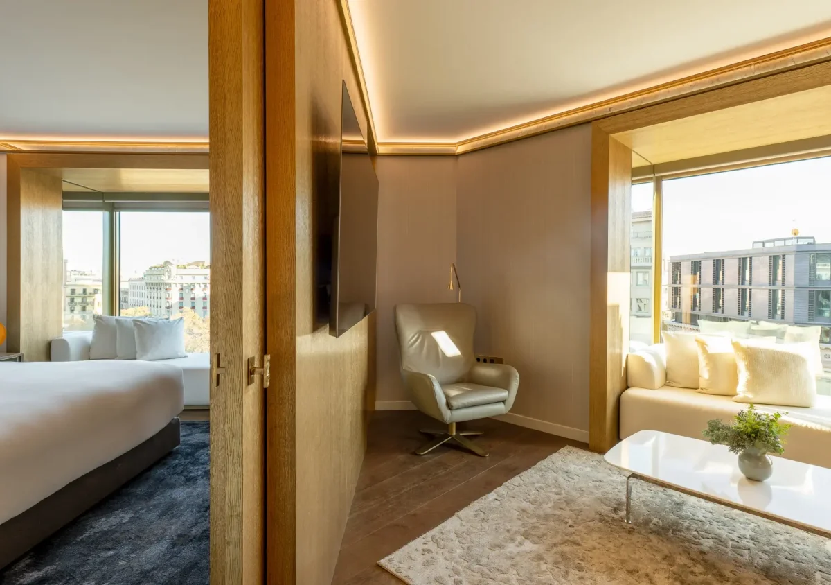 Die Almanac Suite ist eine moderne Hotelsuite mit einem Bett, einem Sessel, einem Sofa, großen Fenstern mit Blick auf die Stadt und einer hölzernen Trennwand mit einem angeschlossenen Fernseher.