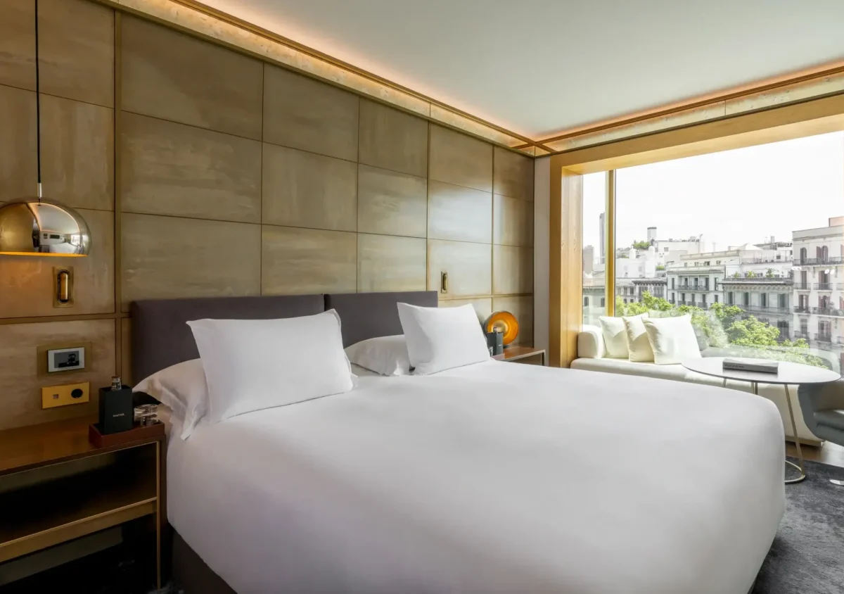 Modernes Hotelzimmer der Almanac Suite mit einem großen Bett, weißer Bettwäsche, Nachttischen, einer goldenen Wandlampe und einem Fensterplatz mit Blick auf die Gebäude der Stadt und die Grünanlagen.