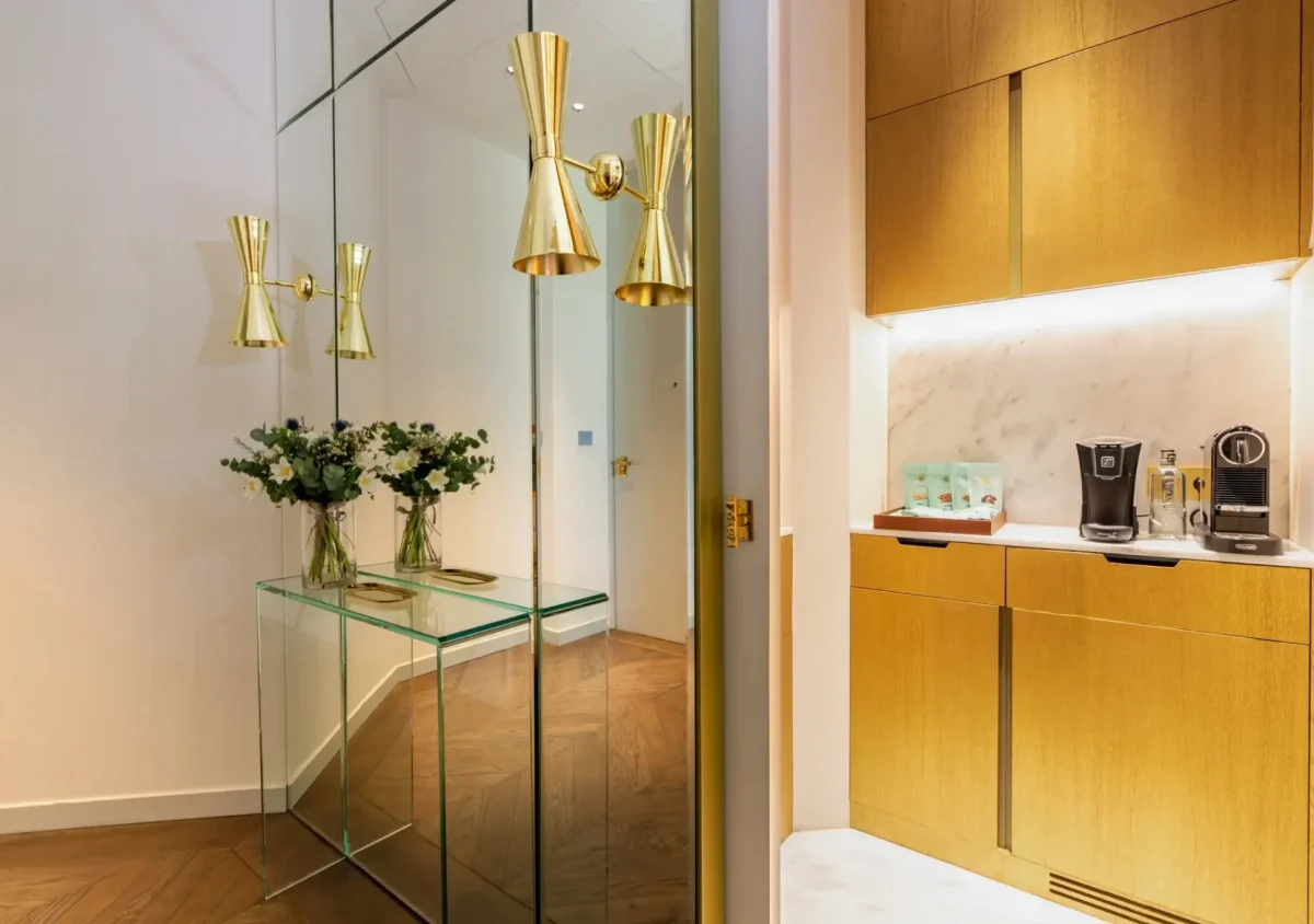 Ein moderner Flur in der Almanac Suite ist mit verspiegelten Wänden, goldenen Leuchten, einem gläsernen Konsolentisch mit Blumenvasen und einer eleganten Küchenzeile mit Kaffeemaschinen und Holzschränken ausgestattet.