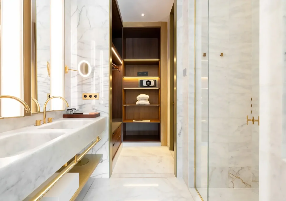 Modernes Badezimmer in der Almanac Suite mit Marmorwänden und -böden, Doppelwaschbecken, großen Spiegeln, goldenen Armaturen und einem begehbaren Kleiderschrank mit Regalen, Handtüchern und einem Safe.