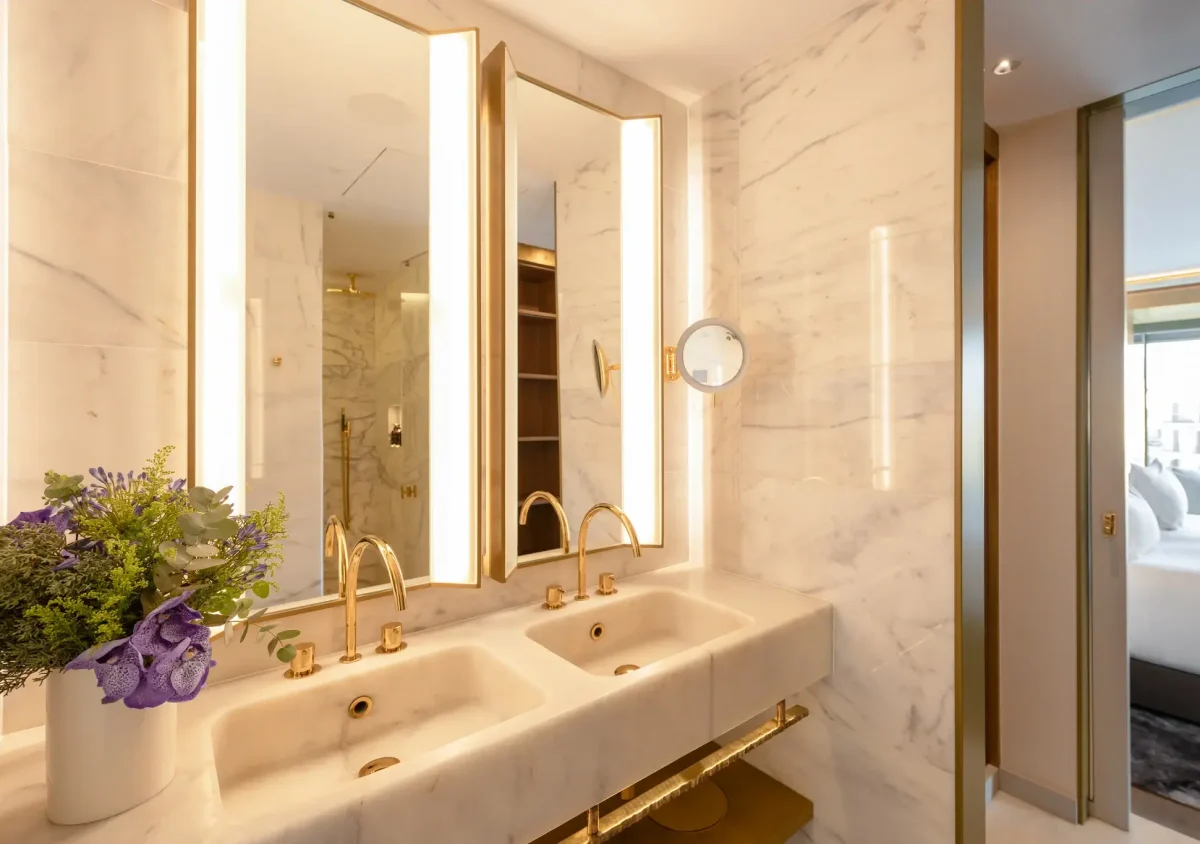 Modernes Badezimmer in der Almanac Suite mit Doppelwaschbecken, goldenen Armaturen, Marmorwänden, großen Spiegeln mit Beleuchtung und einer Vase mit lila Blumen auf dem Tresen. Ein Schlafzimmer ist durch eine offene Tür sichtbar.