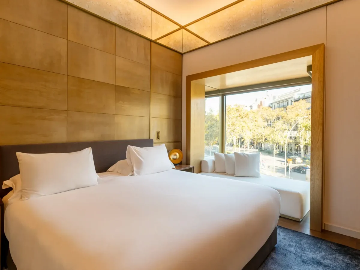 Moderne Almanach-Suite mit einem großen Bett, goldenen Wandpaneelen und einem Fensterplatz mit Blick auf eine belebte, von Bäumen und Gebäuden gesäumte Stadtstraße.