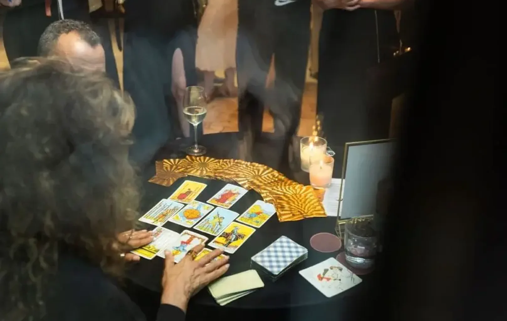 At a Zenith Leo Session, a person with long hair reads tarot cards at a round table adorned with candles and drinks, while an intrigued audience stands around, watching the mystical reading unfold.