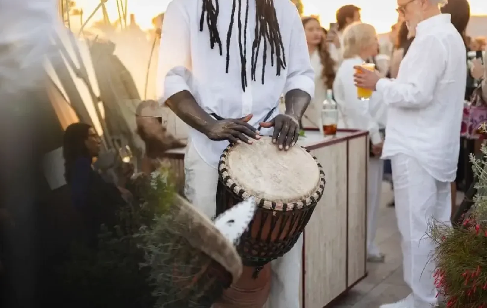 At a Leo Session, a person in white clothing plays a djembe drum at an outdoor gathering, while other attendees—also dressed in white—socialize and hold drinks in the background, celebrating under the day's zenith.