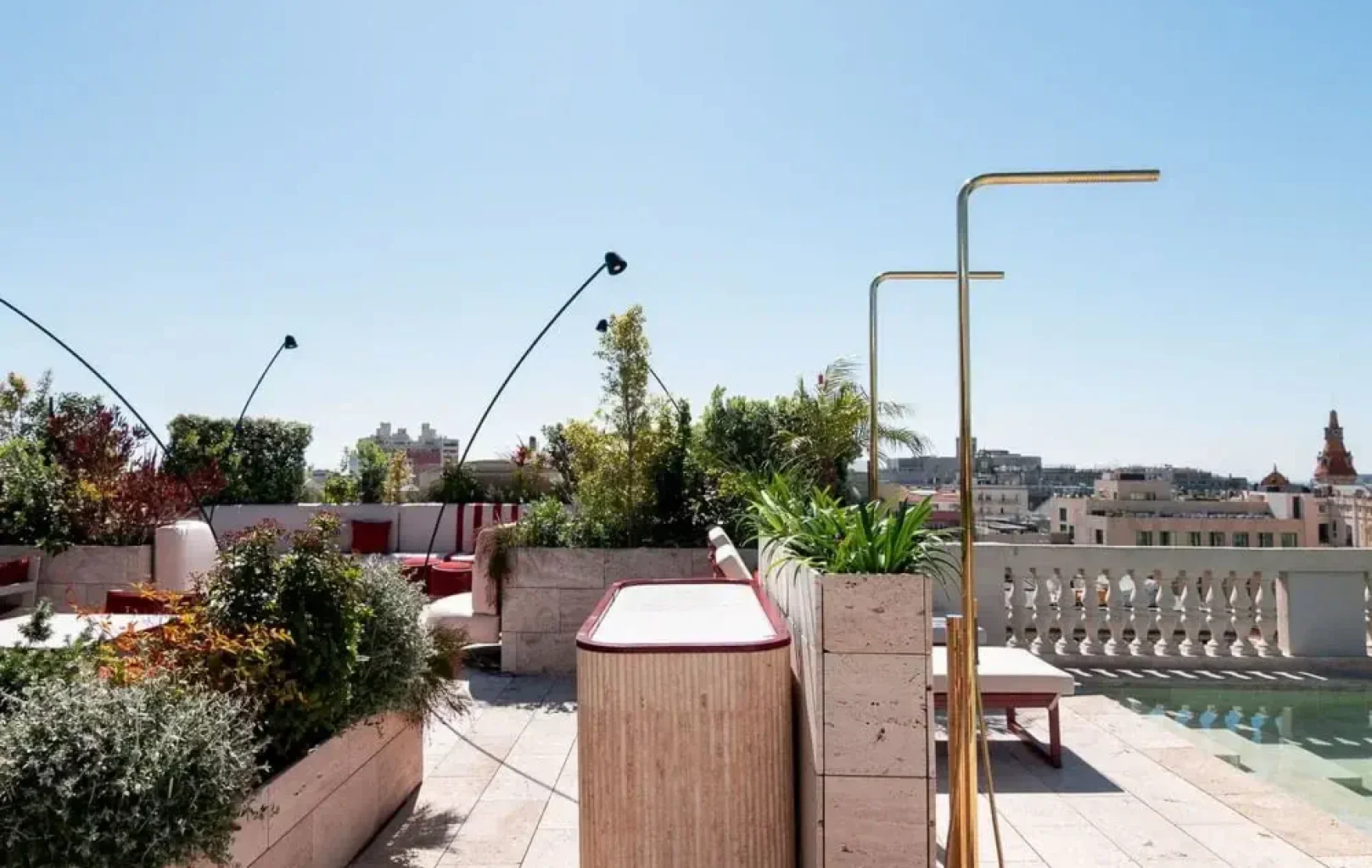 Una terraza en la azotea con asientos empotrados, macetas, una barra de bar, modernas lámparas y edificios urbanos visibles al fondo ofrece una nueva perspectiva bajo un cielo despejado.