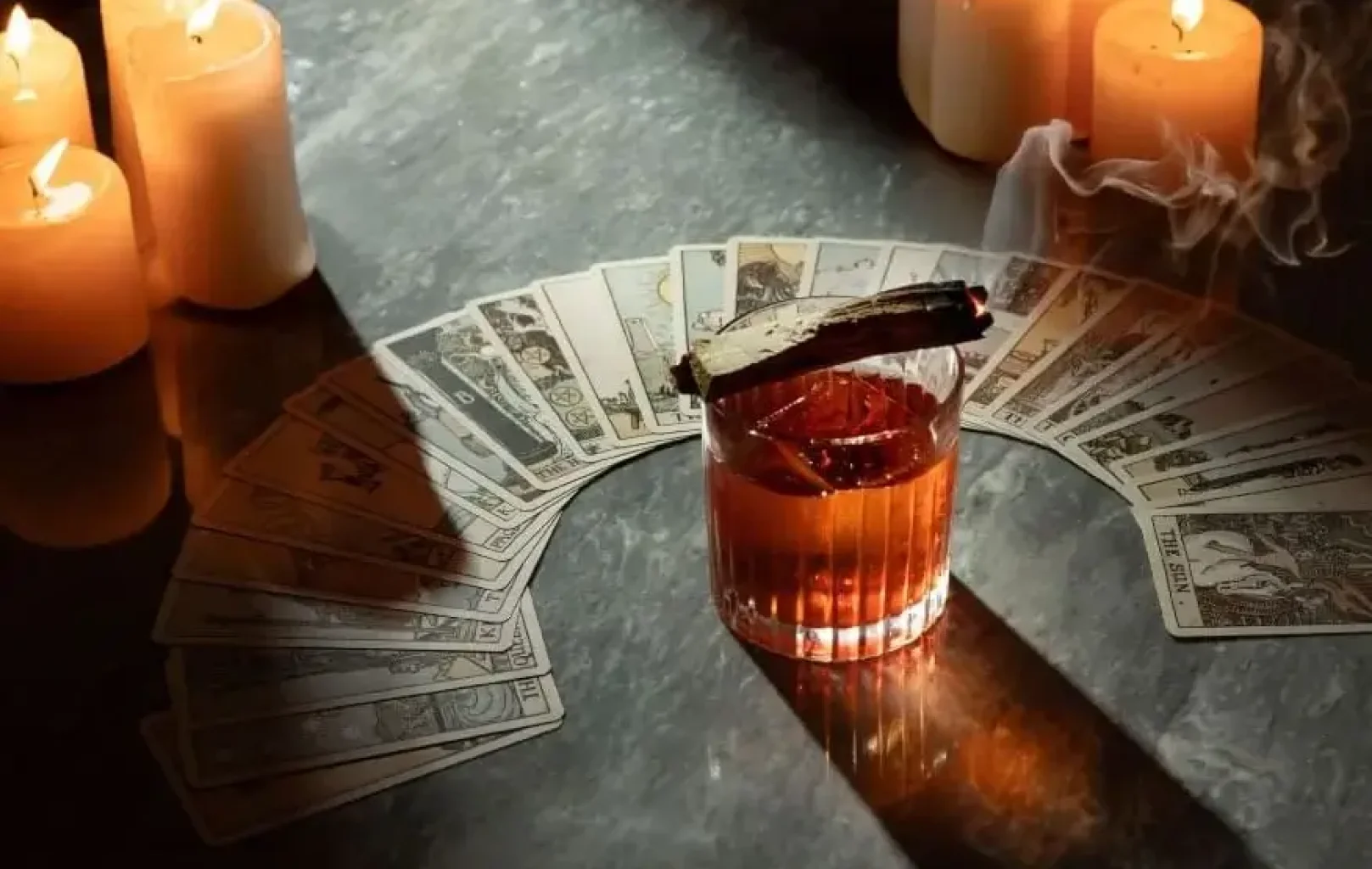 Un vaso de cóctel con una varilla de incienso encendida descansa sobre una tirada de cartas del tarot, ofreciendo una nueva perspectiva mientras las velas encendidas proyectan sombras sobre la oscura superficie.