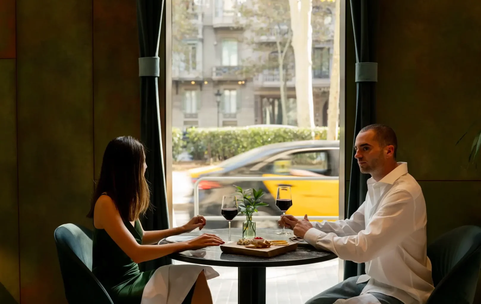 Un home i una dona seuen a una taula rodona amb vi i aperitius davant d'un gran finestral, gaudint de la seva Escapada de Tardor mentre un cotxe groc passa per fora.
