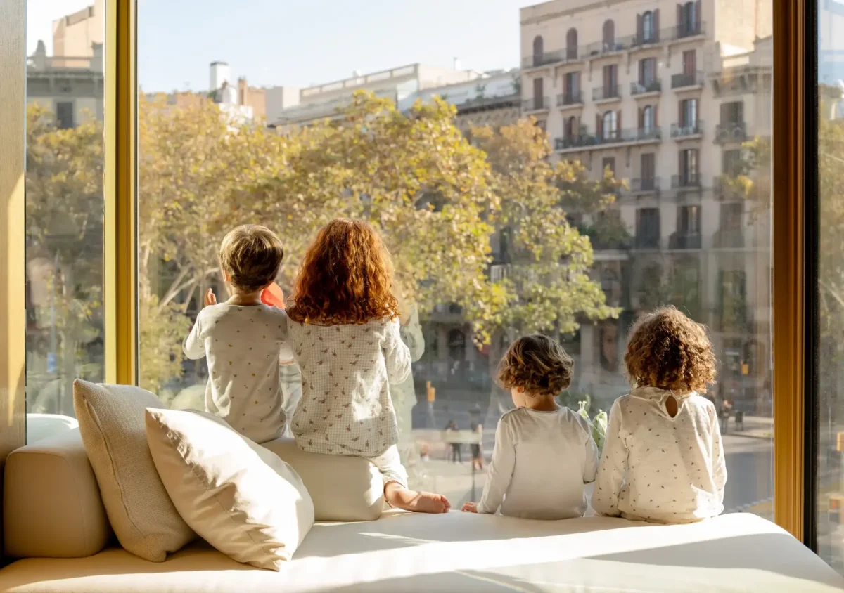 Quatre nens petits en pijama seuen en un seient a la finestra d'un allotjament familiar, mirant un carrer de la ciutat amb arbres i edificis d'apartaments a la llum del dia.