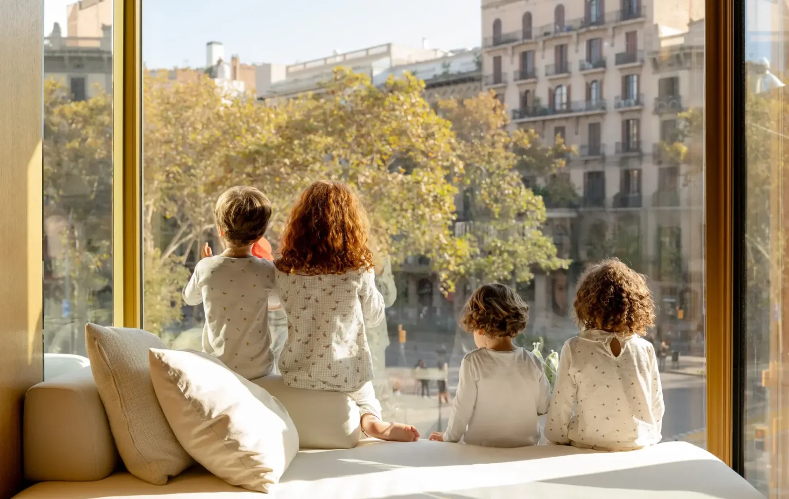 Cuatro niños en pijama se sientan en el alféizar de la ventana de la sala de estar de su familia, contemplando un paisaje urbano salpicado de árboles y edificios de apartamentos en un día soleado.
