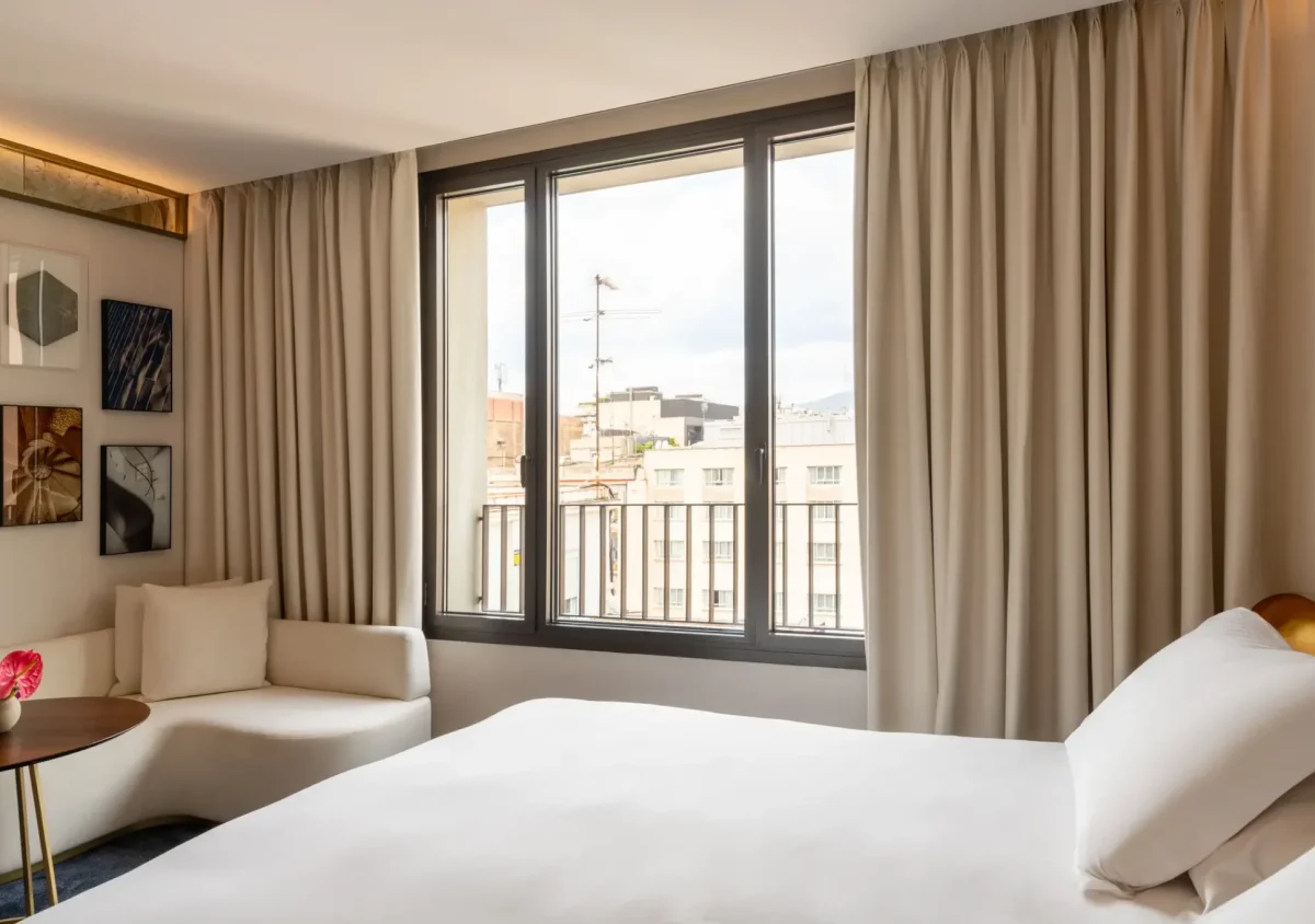 Una sala d'estar familiar moderna comunicada amb un llit blanc, una petita taula rodona amb una flor, un sofĆ en forma de L i grans finestres amb cortines beix amb vistes als edificis de la ciutat.