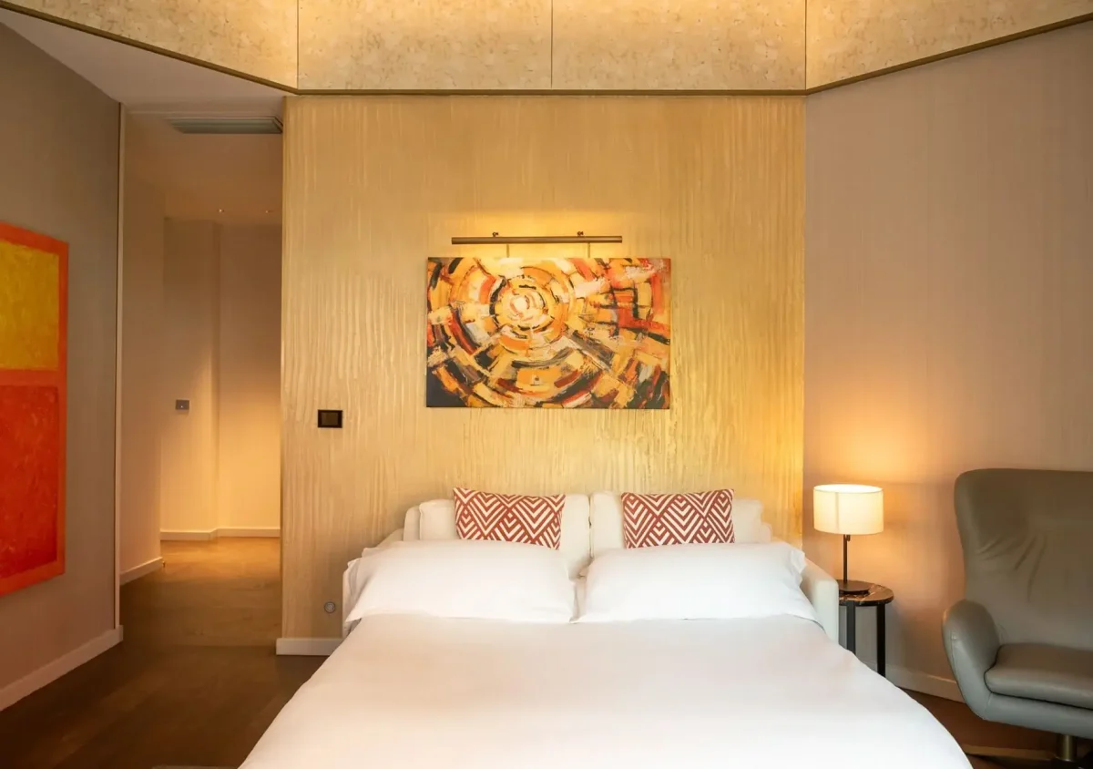 Modernes Schlafzimmer in der Almanac Family Suite mit einem Doppelbett, gemusterten Kissen, einem abstrakten Kunstwerk über dem Kopfteil, einer Nachttischlampe und einem Sessel in einem warm beleuchteten Raum.