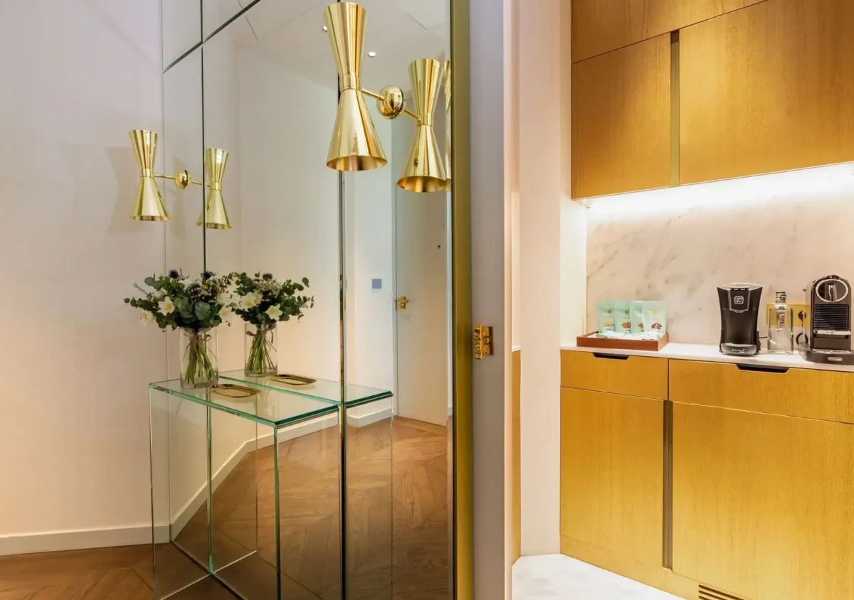 Ein moderner Flur in der Familiensuite ist mit einem gläsernen Konsolentisch, Blumenarrangements, Wandleuchten aus Messing und einer angrenzenden Küchenzeile mit goldenen Schränken und Kaffeemaschinen ausgestattet.