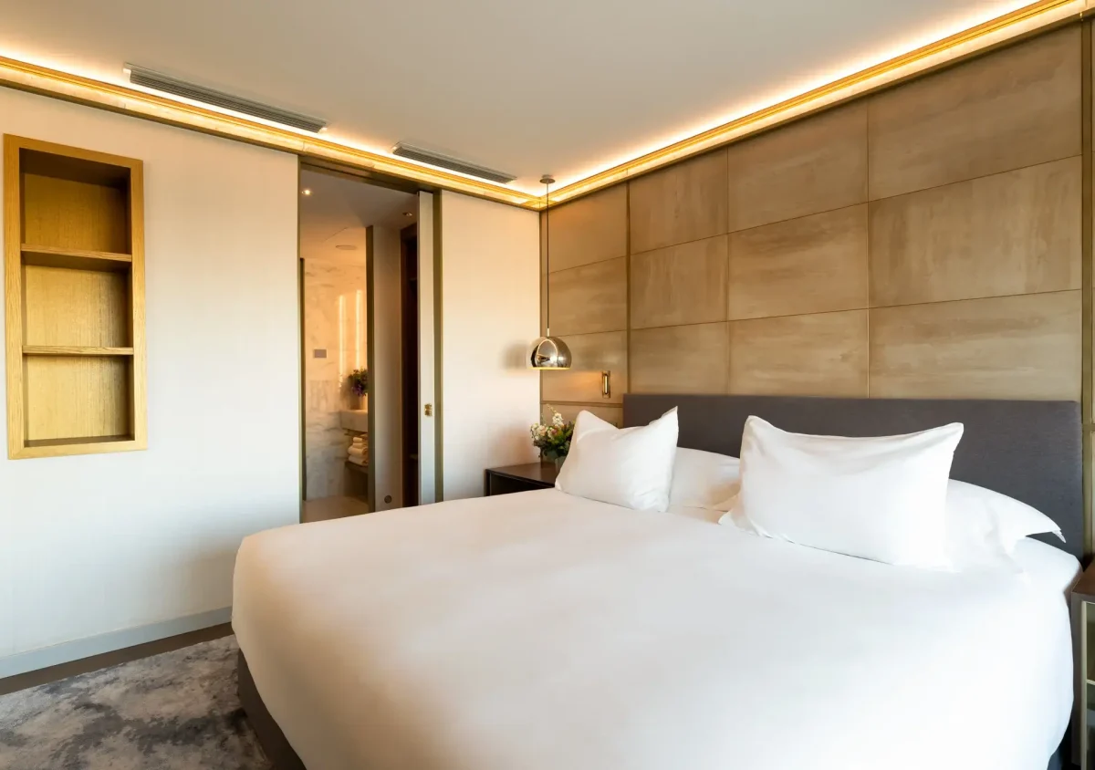 Moderne Almanac Suite mit großem Bett, weißer Bettwäsche, gepolsterter beiger Akzentwand, eingebauten Regalen, Nachttischen und offener Badezimmertür im Hintergrund - perfekt für Gäste, die zwei Schlafzimmer benötigen.