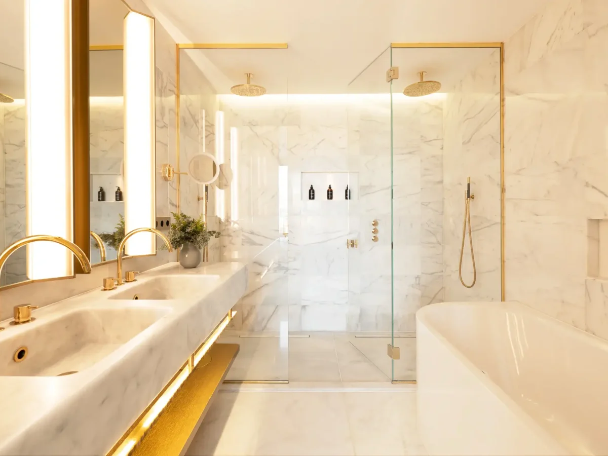 Modernes Badezimmer in der Grand Penthouse Suite mit doppeltem Marmorwaschbecken, goldenen Armaturen, gläserner Dusche, eingebauten Regalen und einer freistehenden Badewanne, alles beleuchtet von hellem Licht.