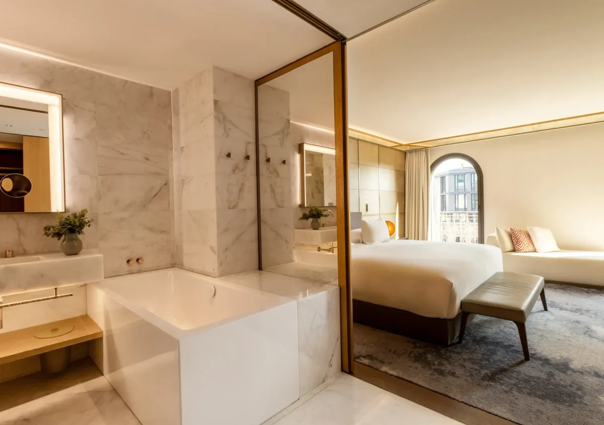 Moderne Junior Suite mit Marmorbad mit Badewanne, großem Spiegel und Glastrennwand, die das Schlafzimmer mit einem bequemen Bett und einer Bank abtrennt.