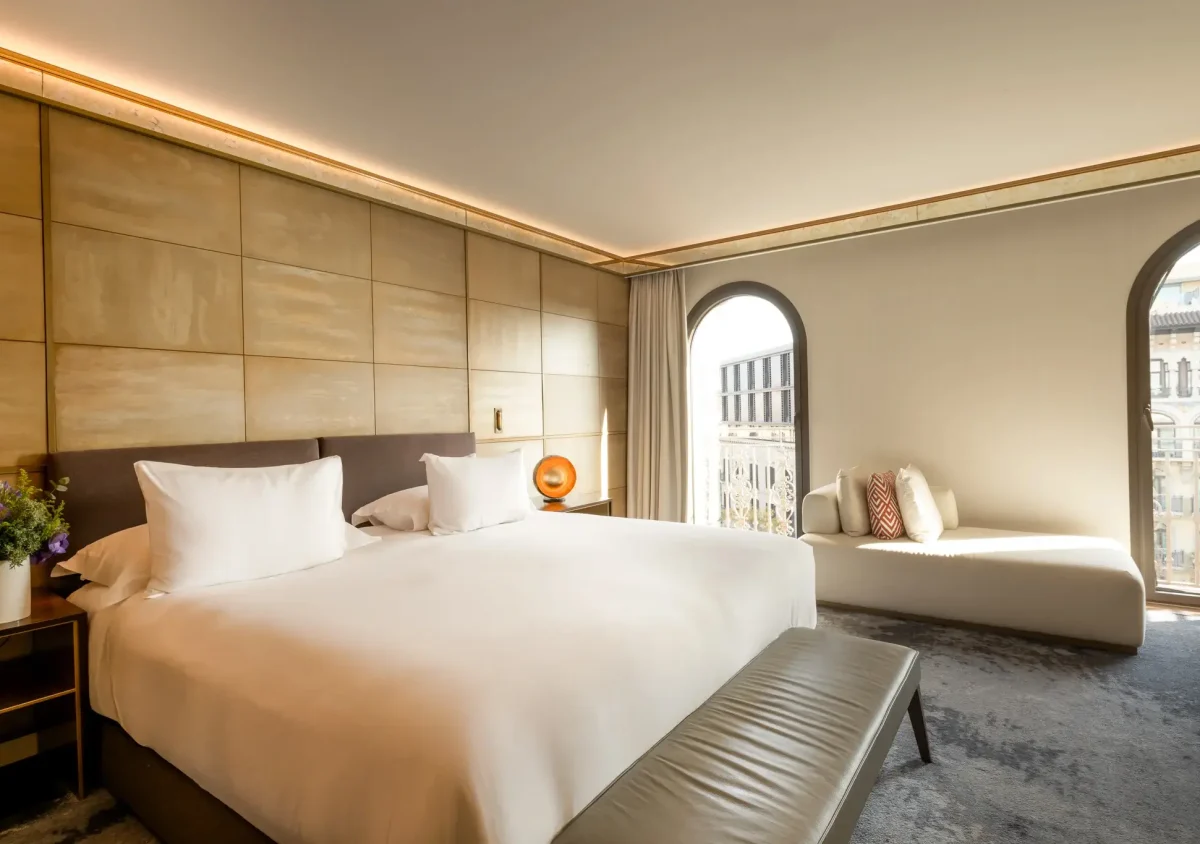 Moderne Junior Suite mit großem Bett, gepolsterter Bank, Beistelltisch mit Blumen und einem Fensterplatz an zwei Bogenfenstern, die den Raum mit natürlichem Licht erfüllen.