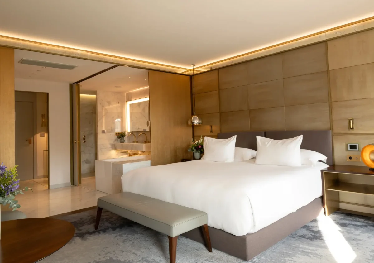 Moderne Junior Suite mit einem großen Bett mit strahlend weißer Bettwäsche, Nachttischen, einer stilvollen Bank am Fußende des Bettes und einem offenen Blick in ein Badezimmer mit luxuriöser Badewanne.