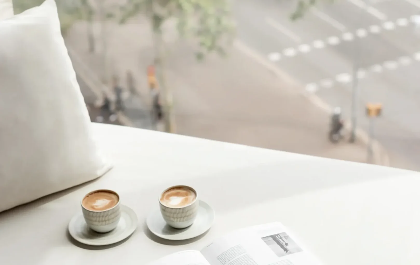 Two cups of coffee and an open book rest on a white surface by a window, offering the perfect autumn escape overlooking a city street lined with trees and traffic.