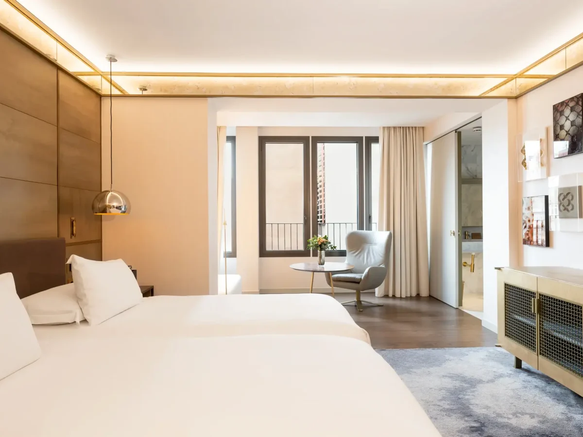 Moderna habitación de hotel con dos camas dobles, una silla y una mesa junto a grandes ventanales, obras de arte en las paredes y un cuarto de baño visible a través de una puerta abierta.