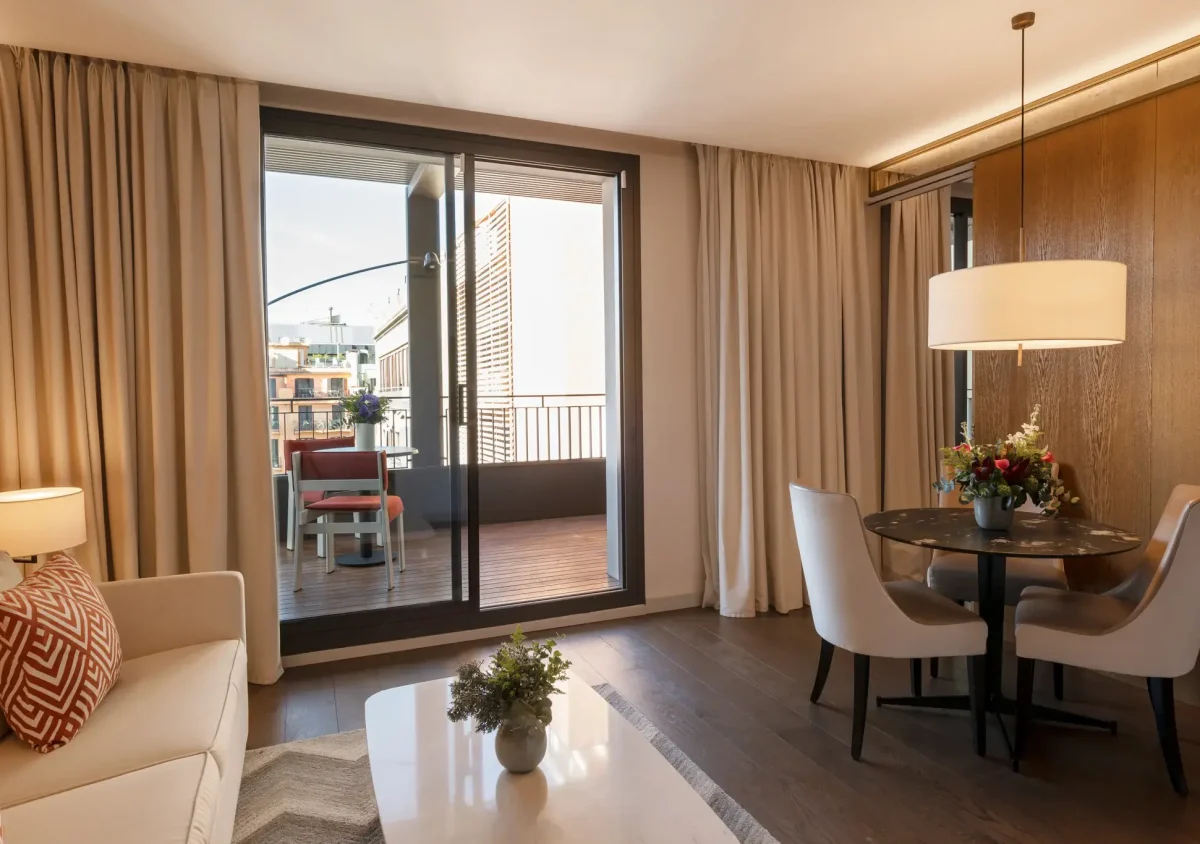 Ein modernes Wohnzimmer mit einem Sofa, einem Couchtisch und einem Essbereich mit großen Glastüren, die sich zu einem Balkon mit Sitzgelegenheiten und Stadtblick öffnen - perfekt, um das Ambiente dieser Terrassensuite zu genießen.