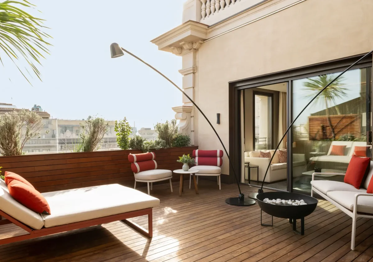 Moderne Terrassensuite im Freien mit Holzterrasse, weißen gepolsterten Lounge-Sesseln und Sofa mit roten Kissen, einer Feuerstelle und Topfpflanzen in der Nähe einer Glastür.