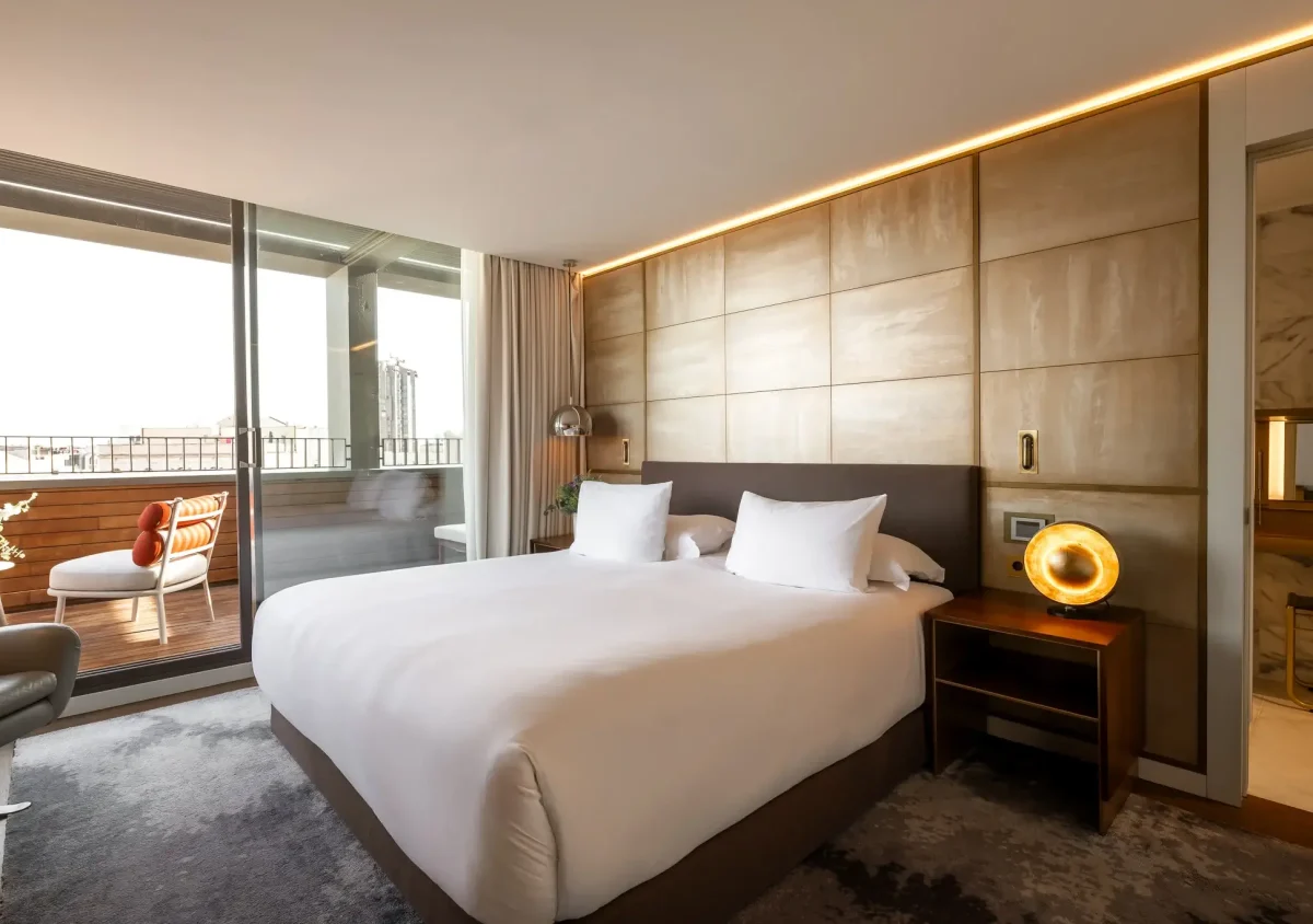 Eine moderne Terrassensuite mit einem großen Bett, zwei weißen Kissen, einem Nachttisch mit Lampe und einem durch Glastüren einsehbaren Balkon mit Sitzgelegenheiten im Freien.