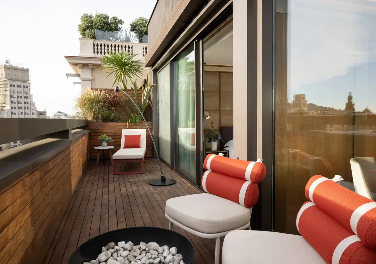 Moderner Terrassensuite-Balkon mit Holzbelag, Loungesesseln mit orangefarbenen Kissen, einer Topfpflanze und einem Blick auf die Stadt durch Glastüren.