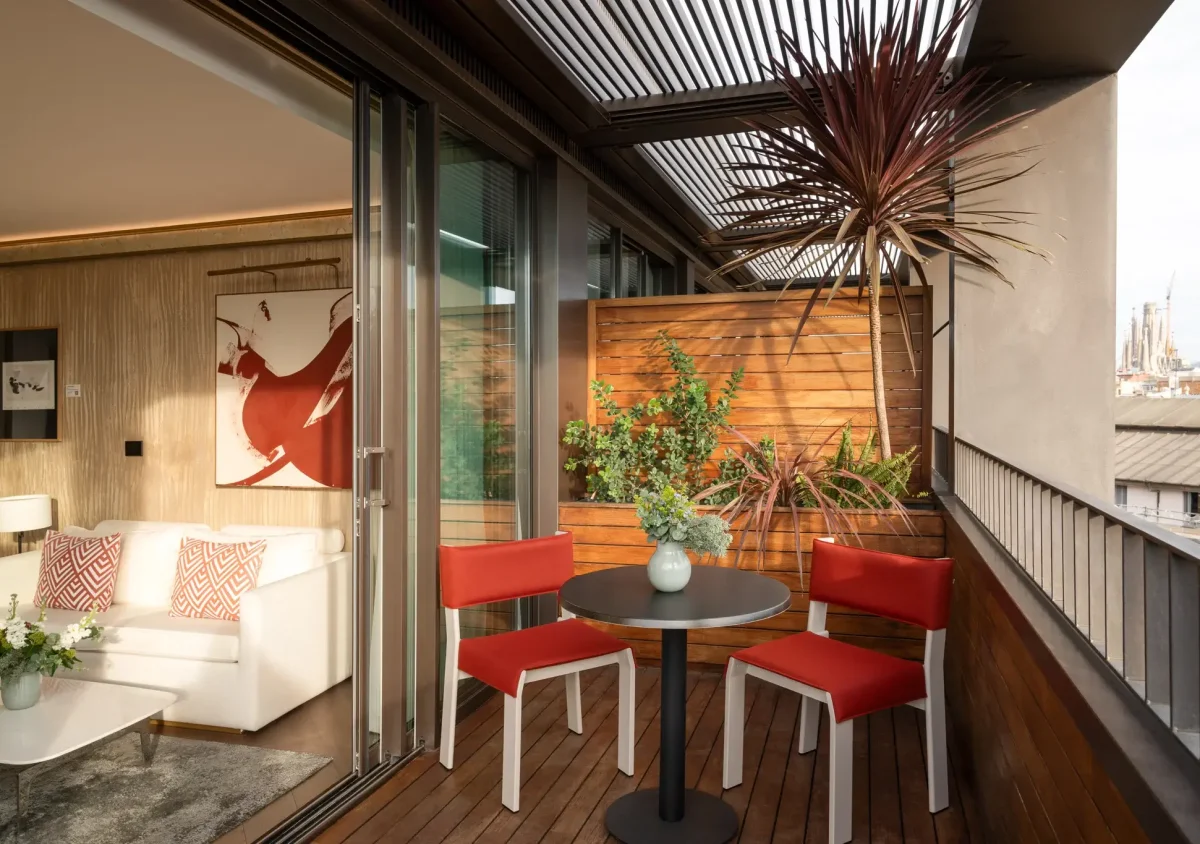 A modern Terrace Suite balcony with a round table, two red chairs, potted plants, and sliding glass doors opening to a living room featuring a white sofa and abstract art.