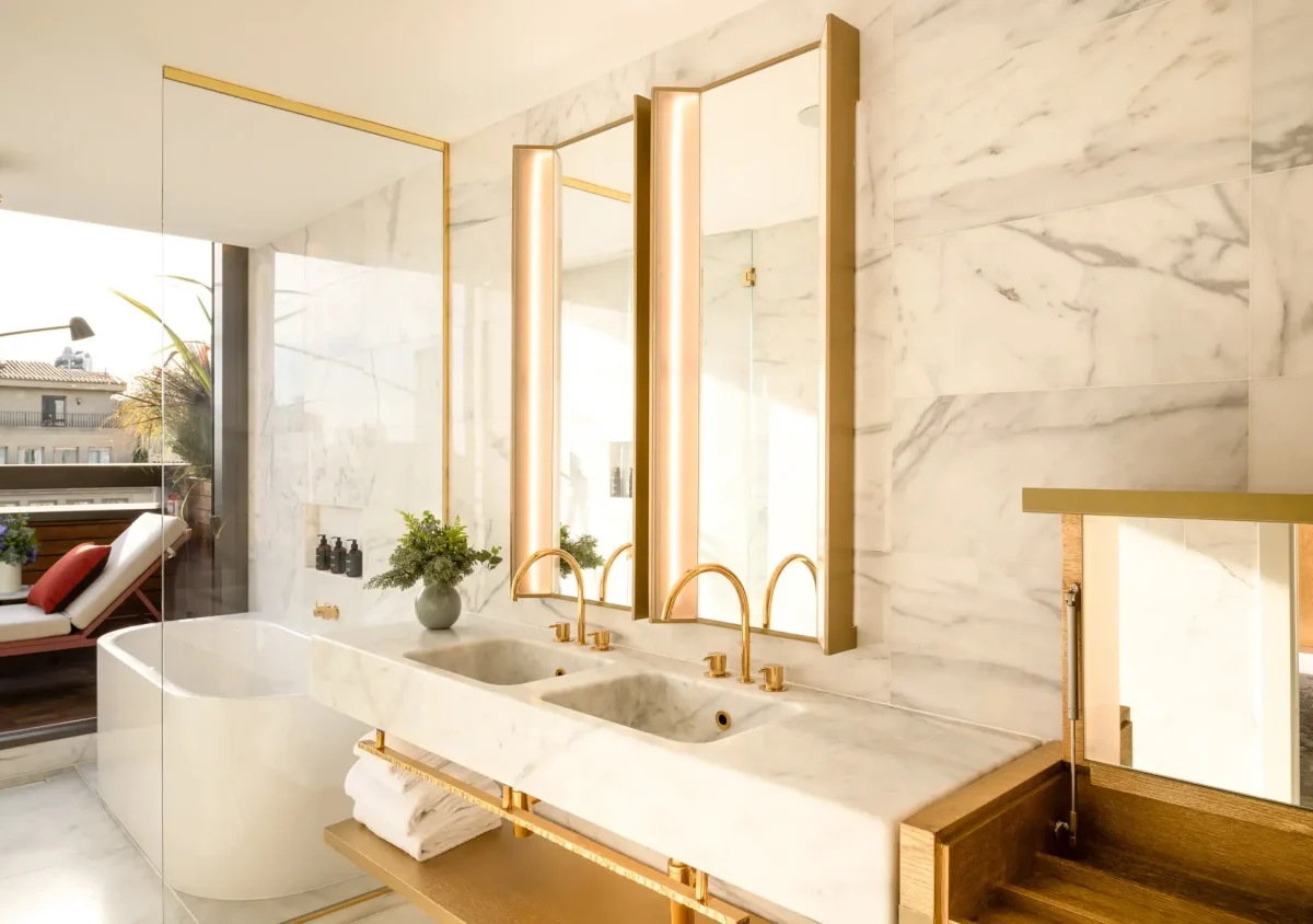 Modern Terrace Suite bathroom with marble walls, double sink vanity with gold fixtures, large mirrors, a freestanding bathtub, and an outdoor seating area visible through glass doors.