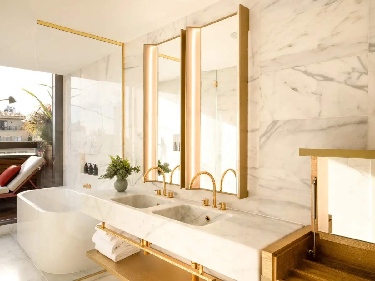 A modern Terrace Suite bathroom with a double marble sink, gold fixtures, a freestanding bathtub, large mirrors, and a view of an outdoor seating area through glass doors.