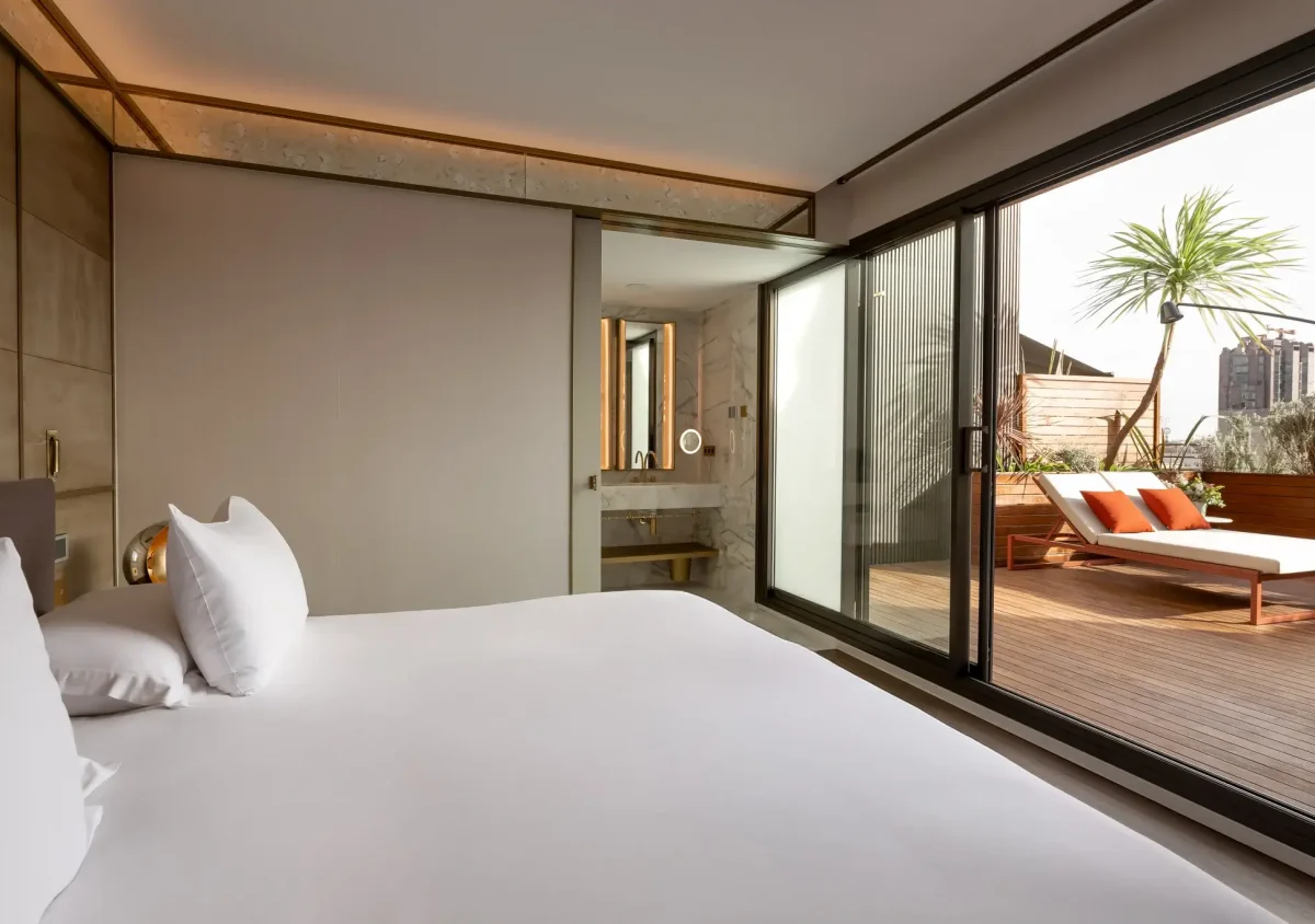 Moderne Terrassensuite mit einem hübsch gemachten Bett und Glasschiebetüren, die zu einer privaten Terrasse mit Liegestühlen, einer Palme und den Gebäuden der Stadt im Hintergrund führen.
