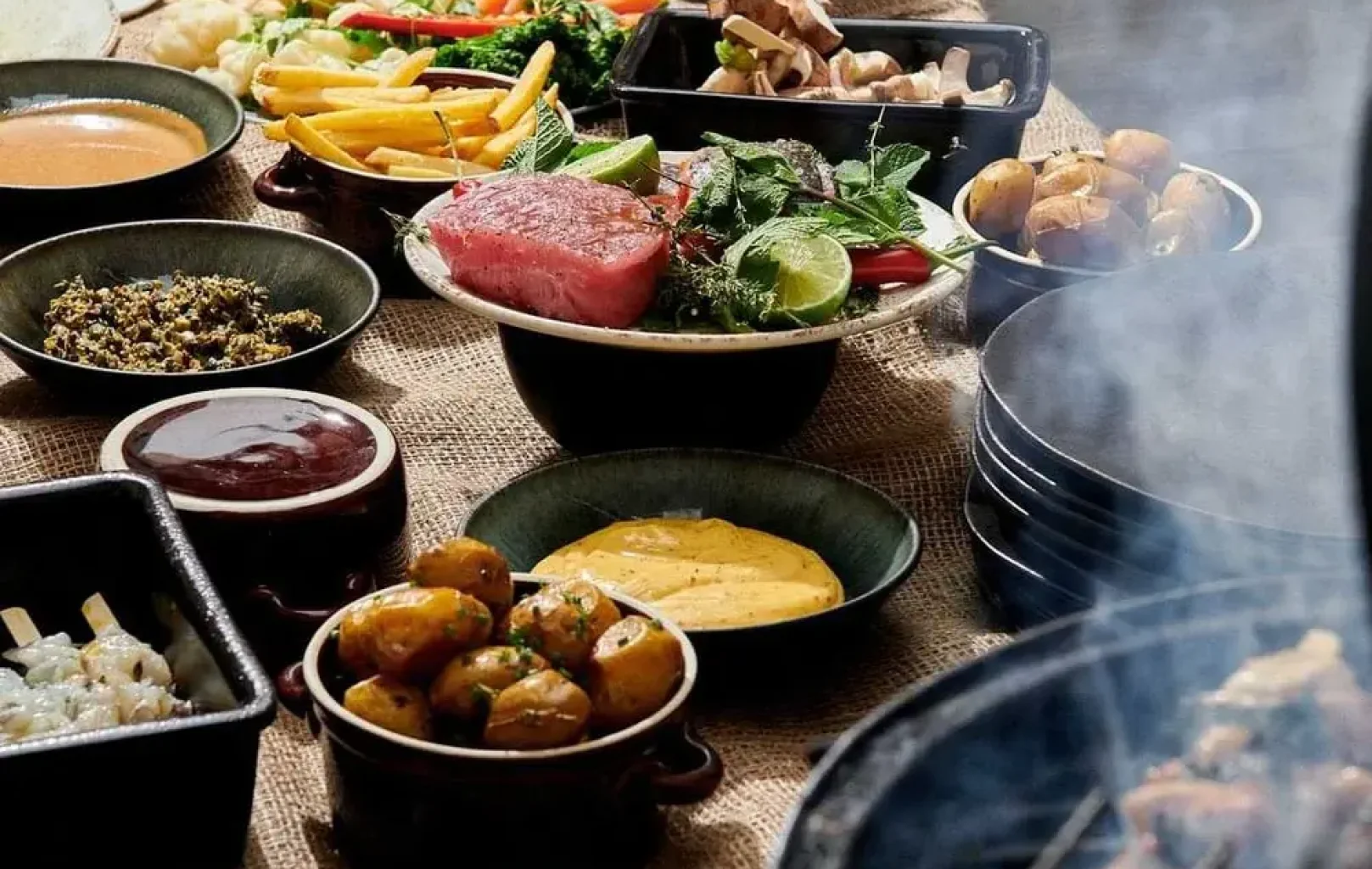 Eine Vielzahl von Gerichten, darunter Soßen, gegrilltes Fleisch, Kartoffeln, Gemüse und Grünzeug, stehen auf einem Tisch neben einem Grill, auf dem das Essen brutzelt - perfekt für ein leckeres BBQ-Lunch.