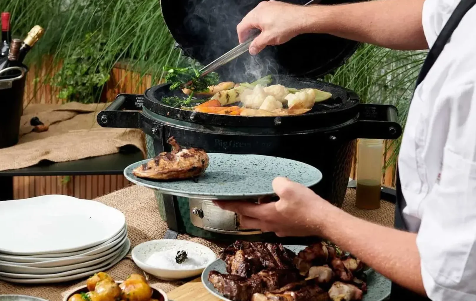 Eine Person grillt Gemüse und Fleisch auf einem Holzkohlegrill für ein BBQ-Mittagessen, wobei verschiedene gekochte Gerichte und Teller auf einem Tisch in der Nähe angeordnet sind.