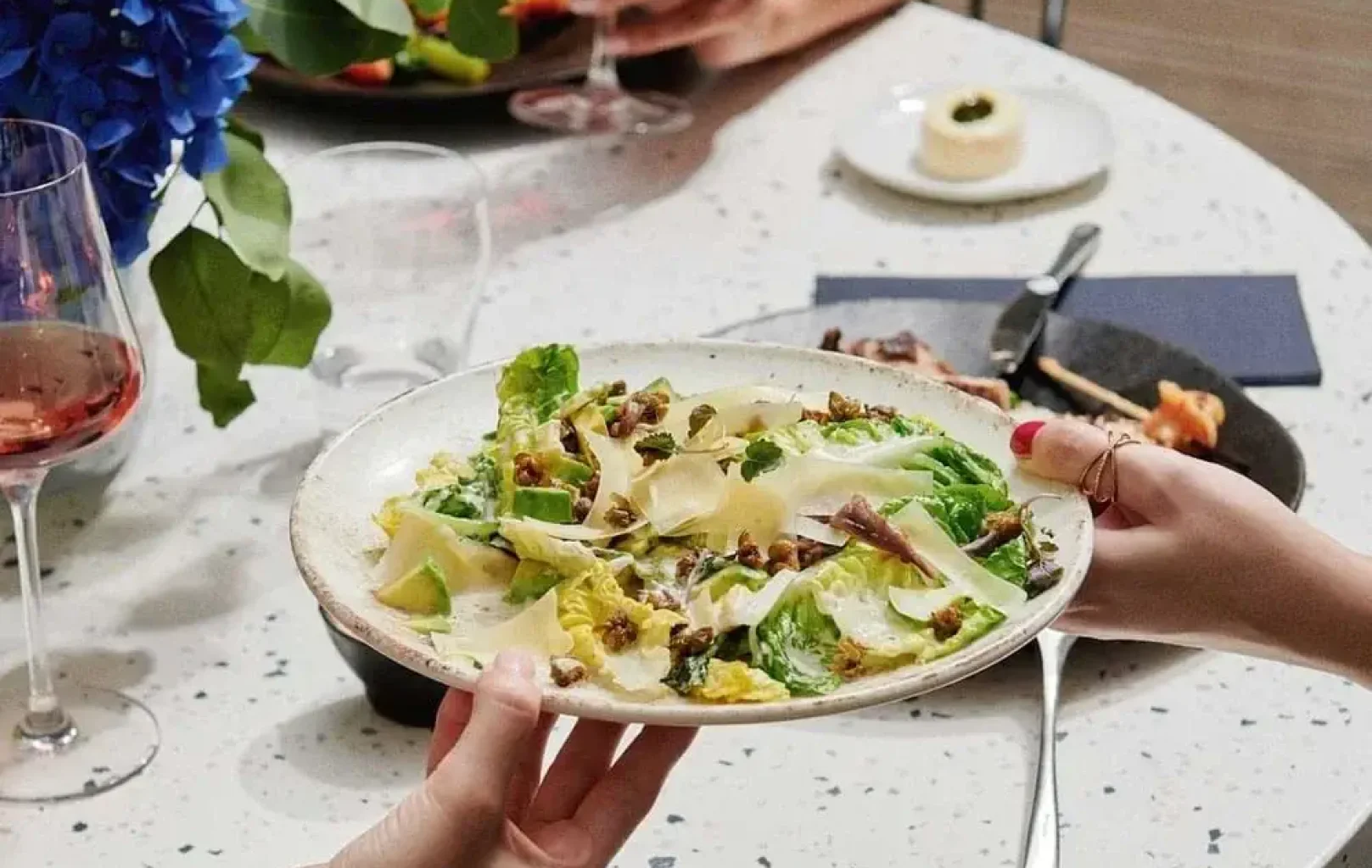 Person mit einem Teller Caesar-Salat mit Salat, Parmesan, Croutons und Dressing an einem Esstisch, der für ein Grill-Mittagessen mit Wein und anderen köstlichen Speisen gedeckt ist.