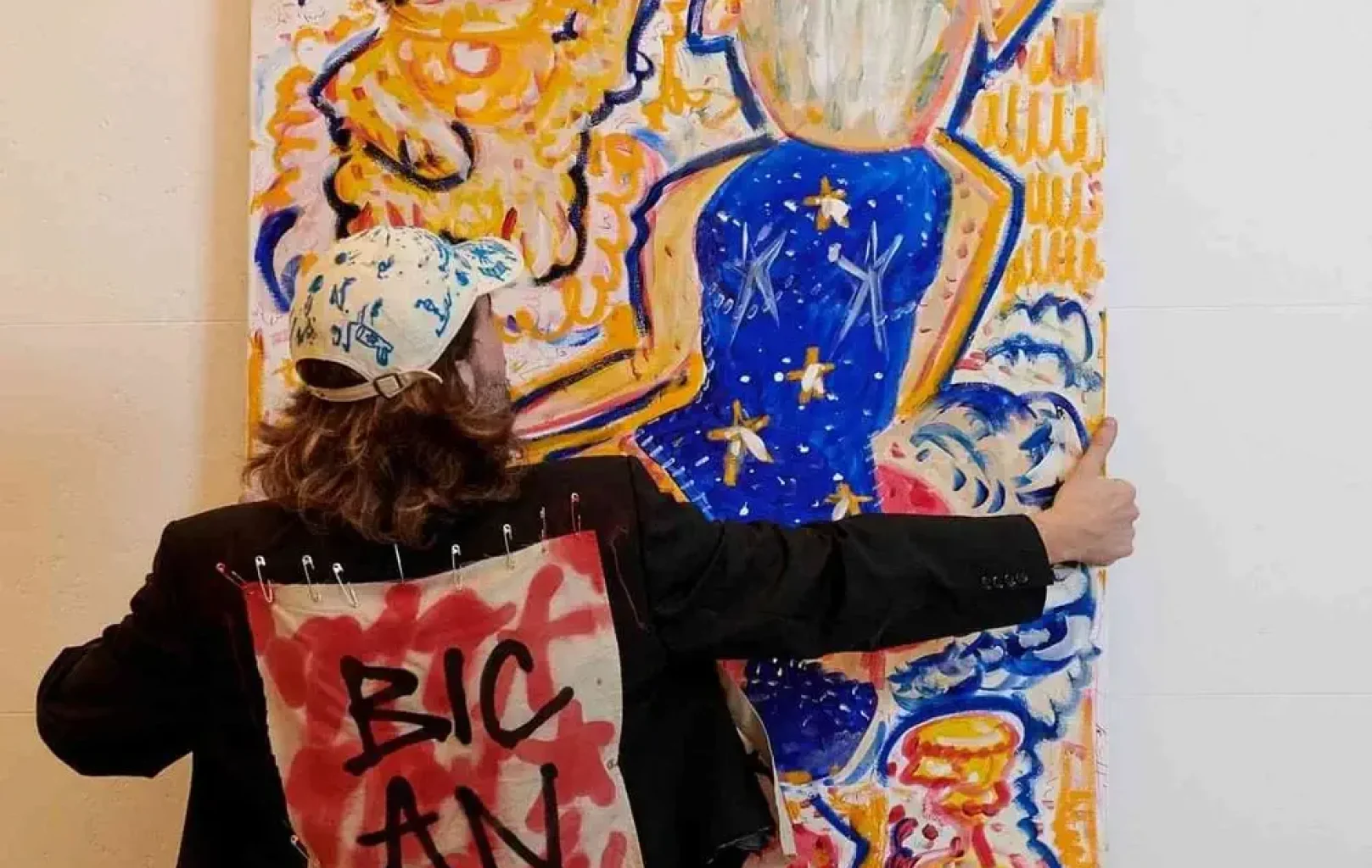 A person wearing a cap and a jacket with "BIC AN" spray-painted on the back holds up a colorful, abstract painting against a wall, capturing the creative spirit found on Signature Tours.