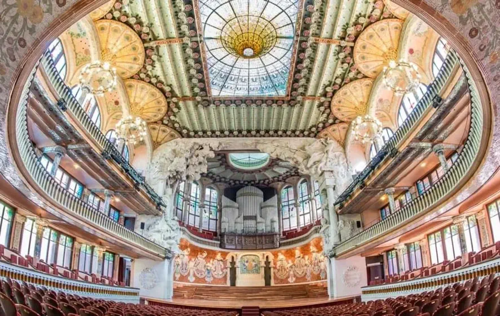 Interior ornamentat d'una sala de concerts d'estil Modernisme Català, amb un sostre decoratiu, una claraboia de vitralls, balcons, parets esculpides i un escenari buit davant de fileres de seients vermells, esperant la seva propera invitació privada.