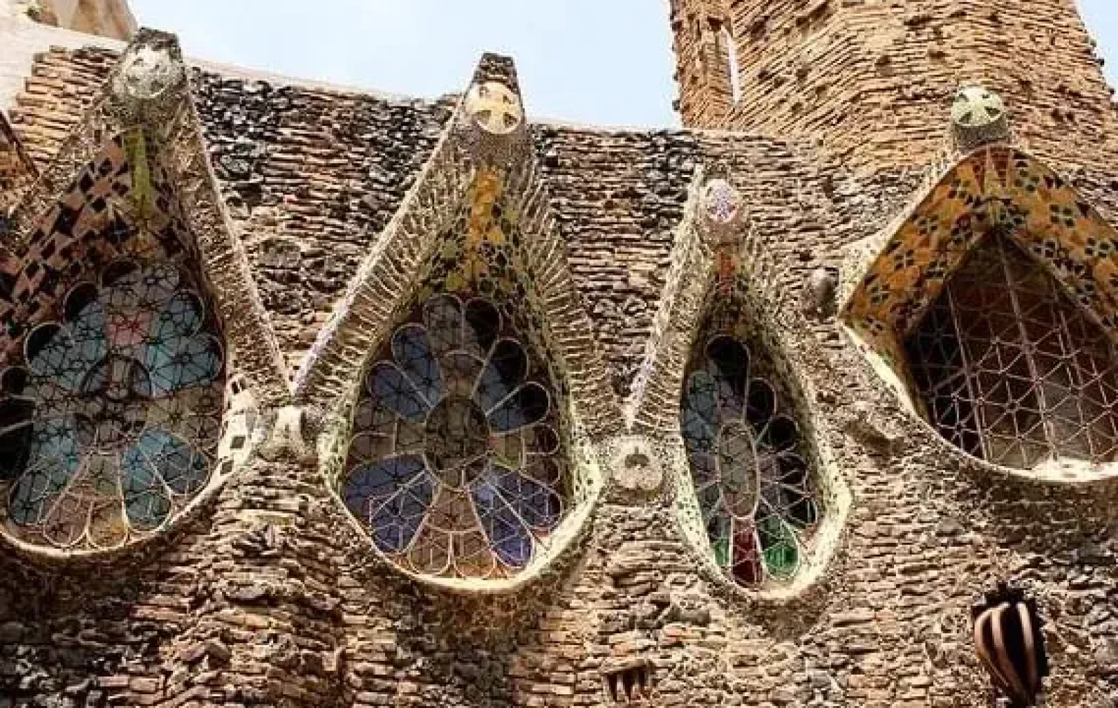 Quatre vitralls amb detalls de mosaic encastats en un mur de pedra texturat, exemplificant el Modernisme Català, part d'un edifici històric amb una torre al fons.