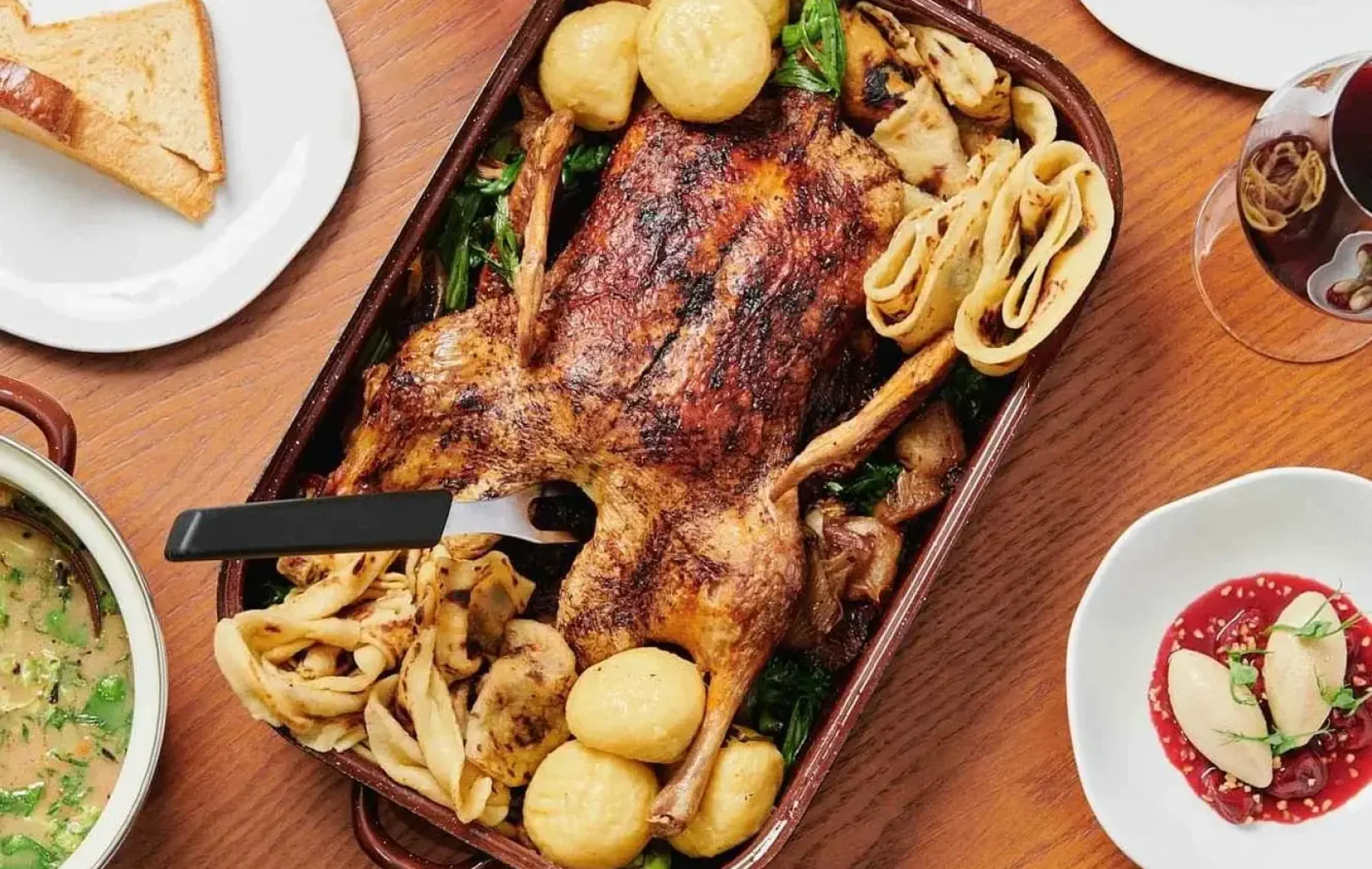Eine gebratene Ente mit Kartoffeln und Gemüse wird in einer Auflaufform serviert, umgeben von Tellern mit Brot, Soße, Rotkohl, Suppe und Gläsern mit Rotwein auf einem Holztisch.