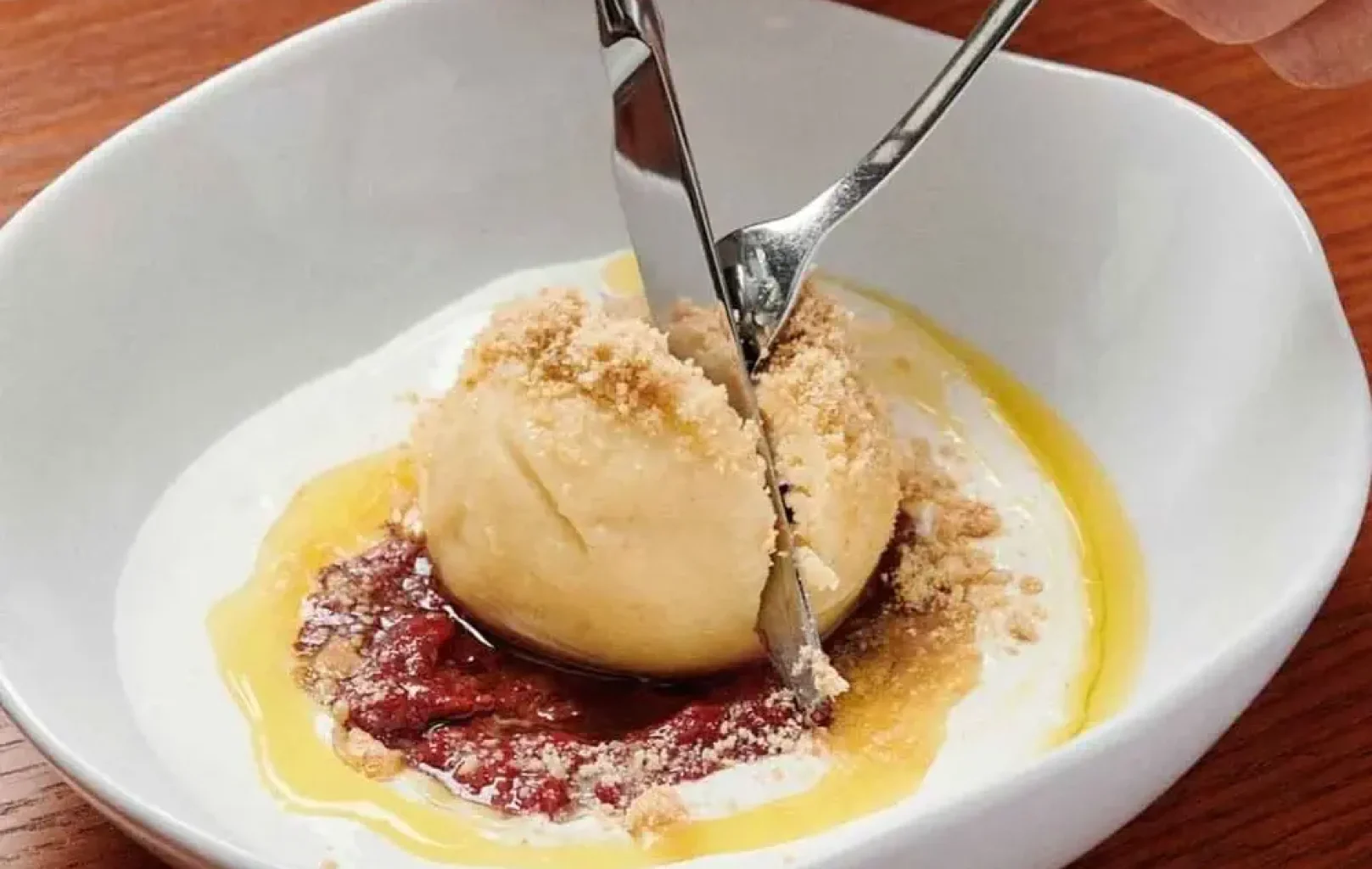 Eine Person schneidet in eine Kugel Eis mit Krümeln, die mit Soße und Sahne in einer weißen Schale auf einem Holztisch serviert wird.