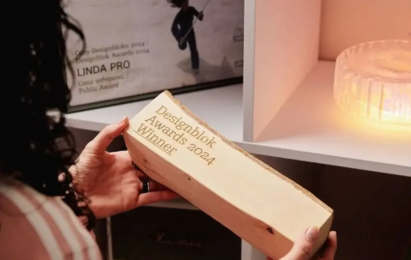 Eine Person hält einen Holzblock mit der Gravur "Designblok Awards 2024 Winner" in einem Raum mit Regalen und Dekorationsgegenständen, die den innovativen Designgeist von Linda Pro zeigen.