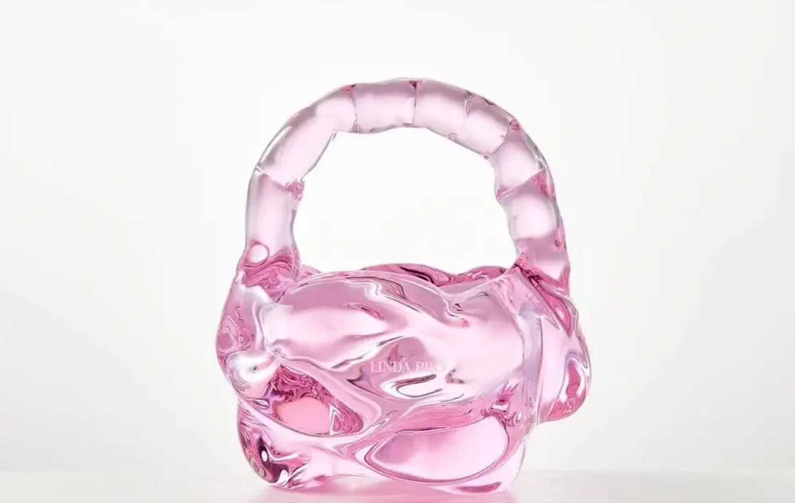 Diese skulpturale Handtasche aus rosafarbenem Glas mit abgerundetem Henkel besticht durch ihr auffälliges Linda Pro-Design vor einem schlichten weißen Hintergrund.